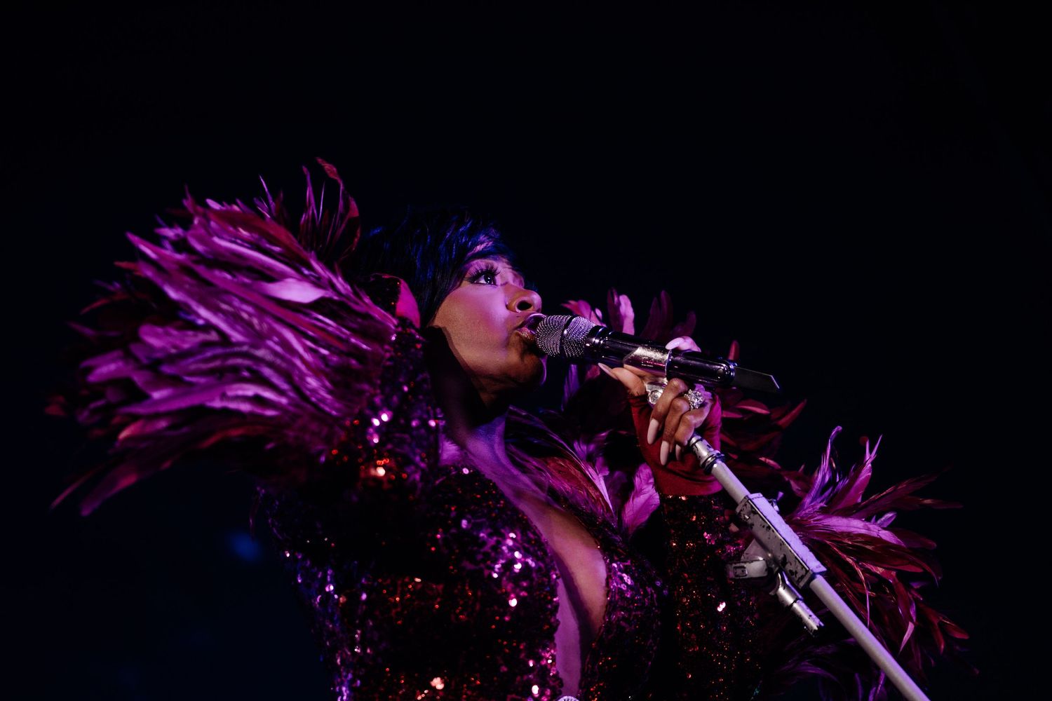 A performer in a sparkling purple outfit with feathered shoulders sings passionately into a microphone on stage.
