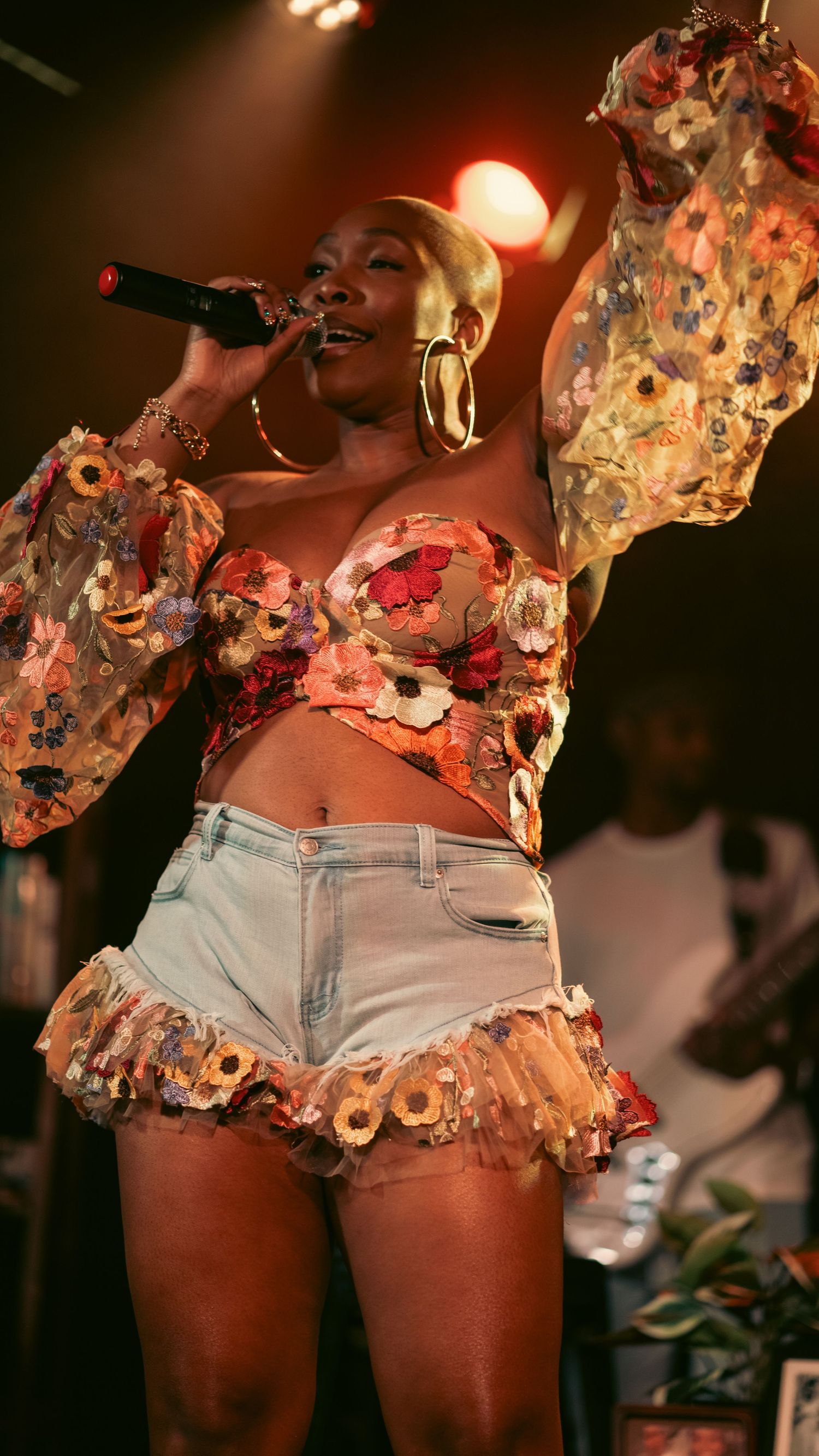 A woman performs on stage wearing a floral bustier top and denim shorts, singing into a microphone under warm stage lights.