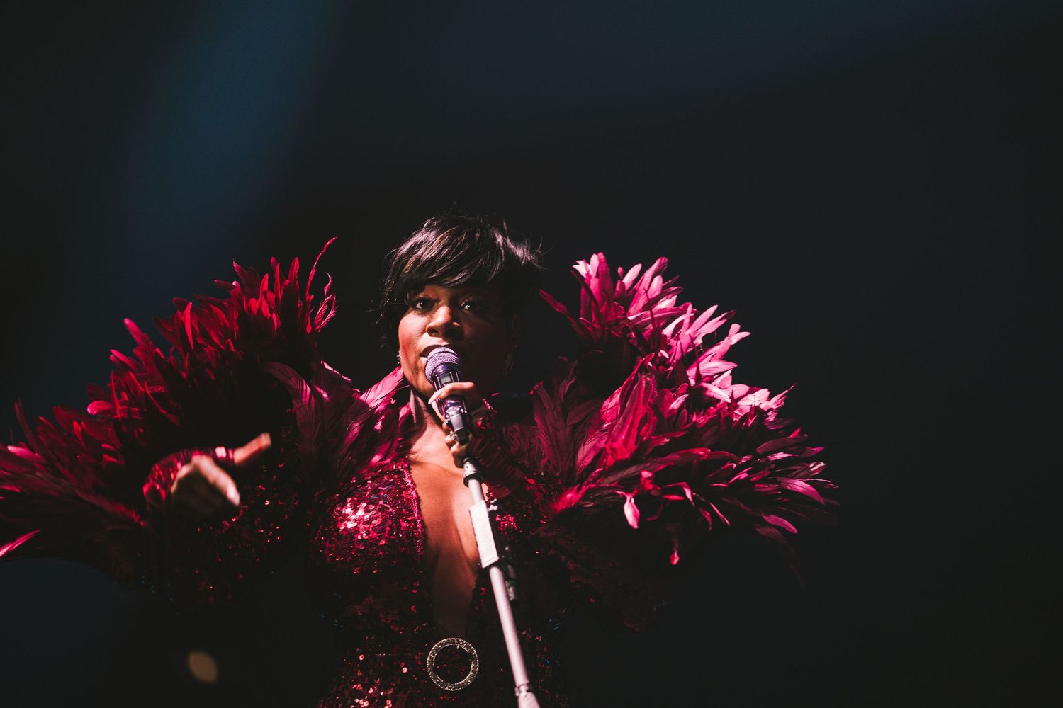 A performer sings on stage wearing a stunning crimson feathered costume under dramatic dark lighting.