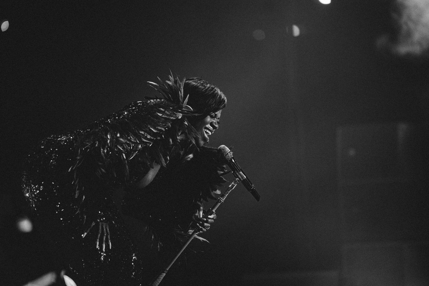 Black and white photo of a performer in feathered costume singing into a microphone on a dark stage.