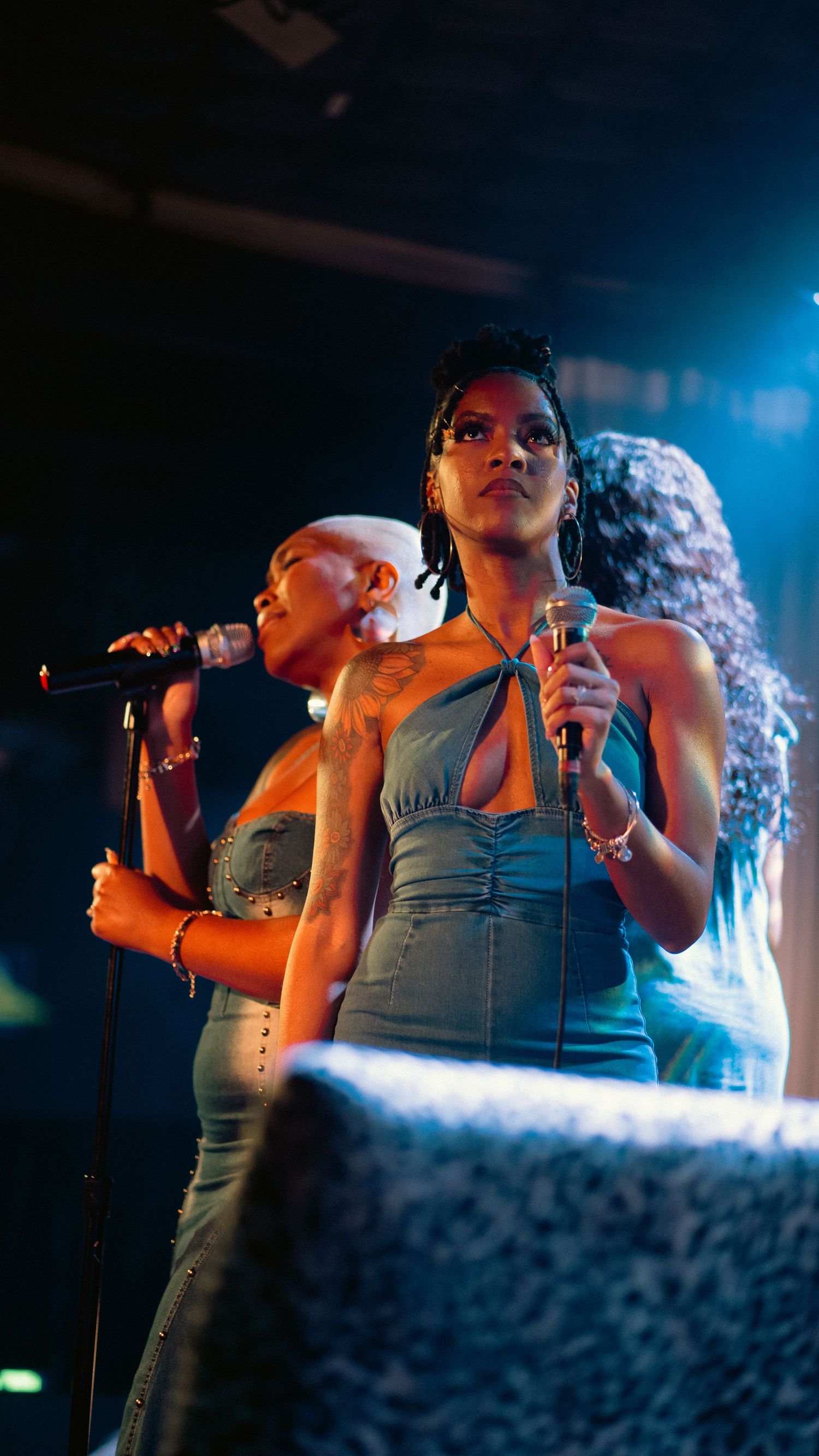 Two female singers in teal dresses perform on stage with microphones under blue lights.