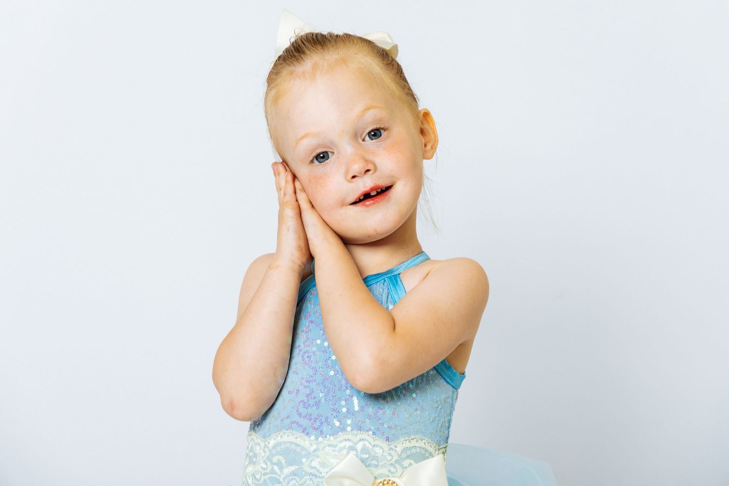 Adorable little girl in blue ballet leotard posing with hands on cheeks, smiling against white background.