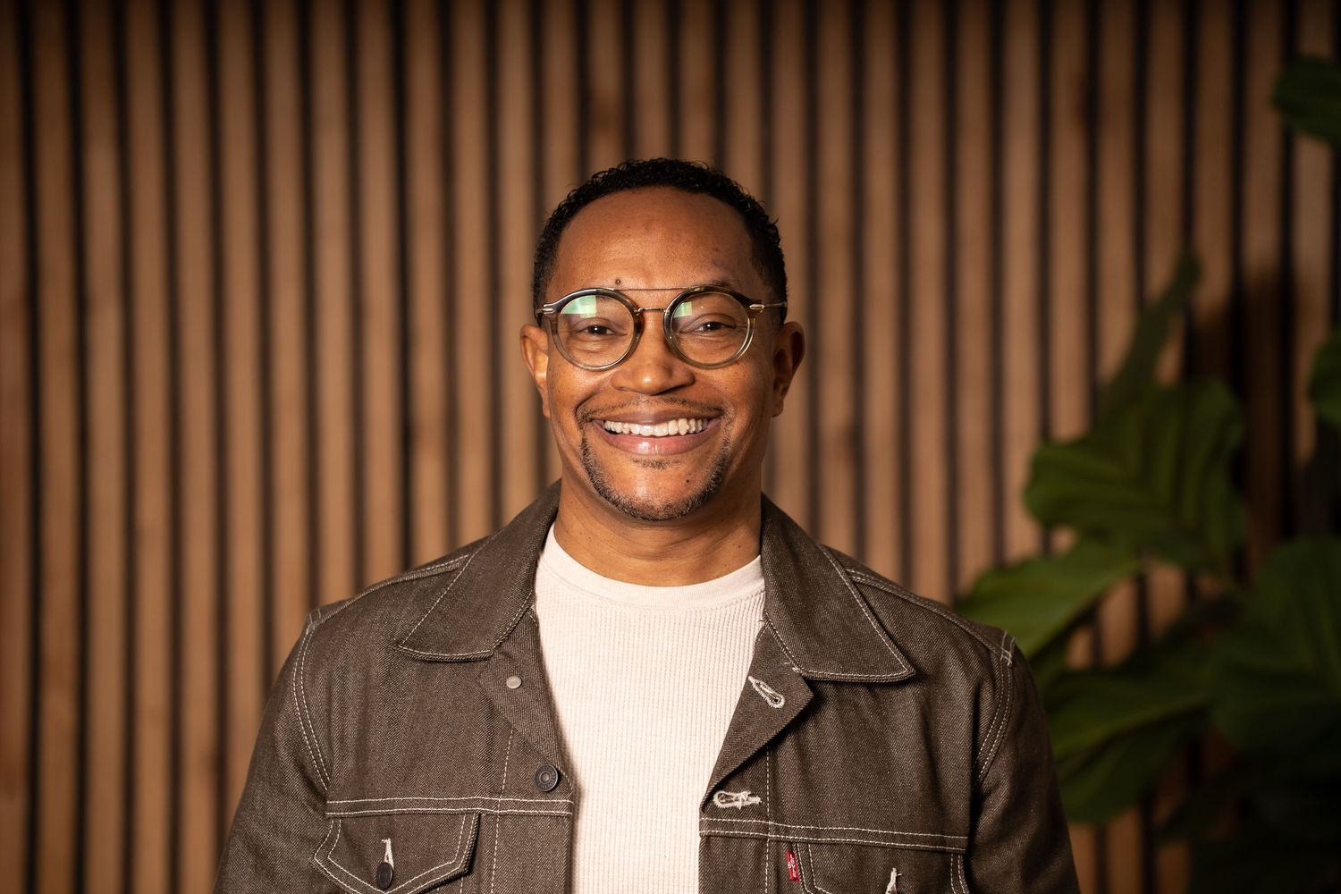 A smiling person wearing glasses and a brown denim jacket poses against a wooden slat wall with warm lighting.