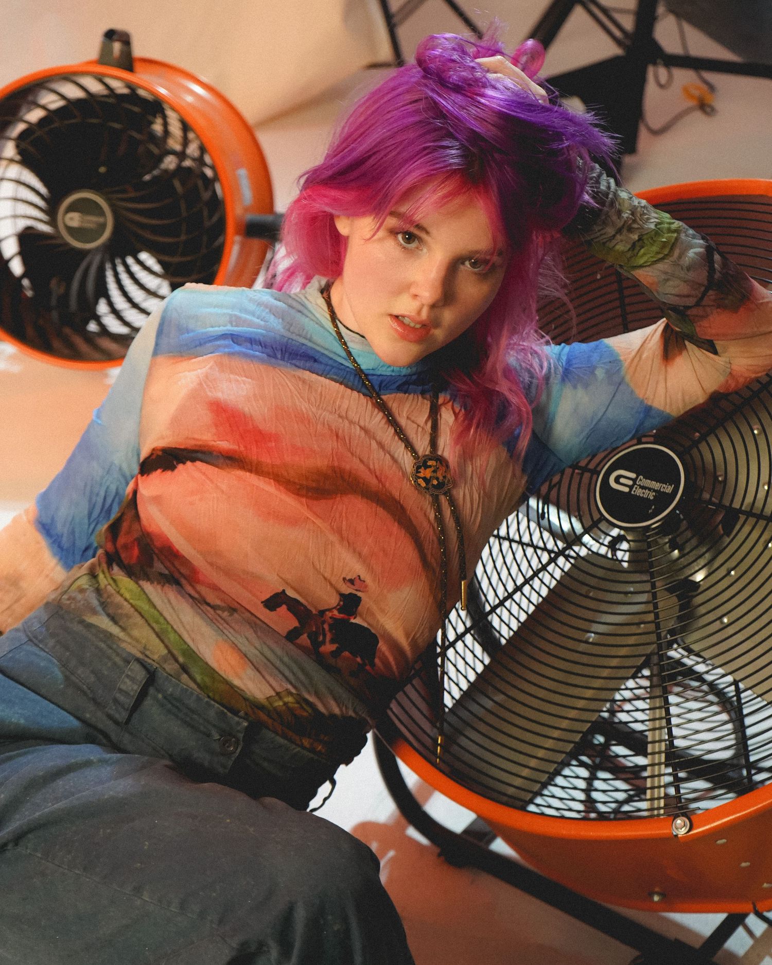 Model with pink hair poses with vintage orange fans wearing denim and colorful jacket.