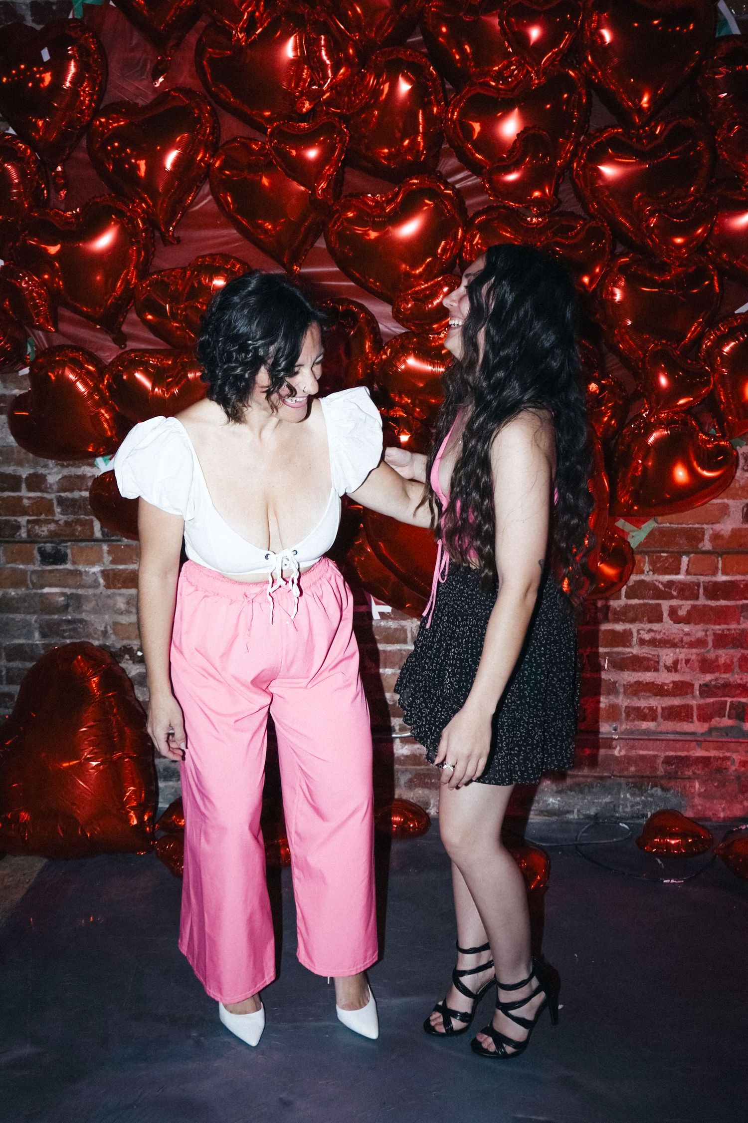 Two figures pose against a brick wall with red heart balloons in a festive Valentine's Day scene.