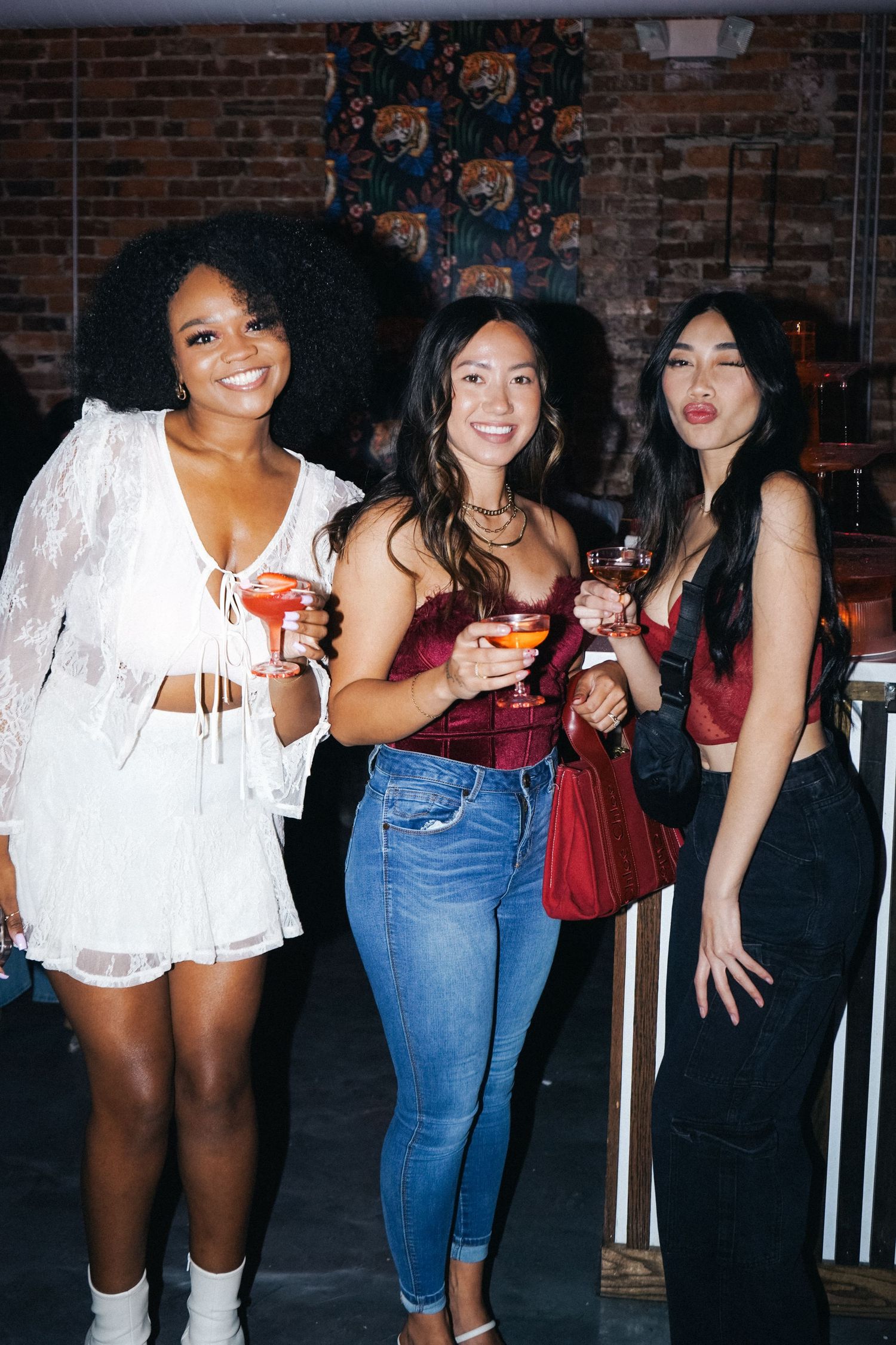 Three friends holding cocktails and smiling at casual party with brick wall background.