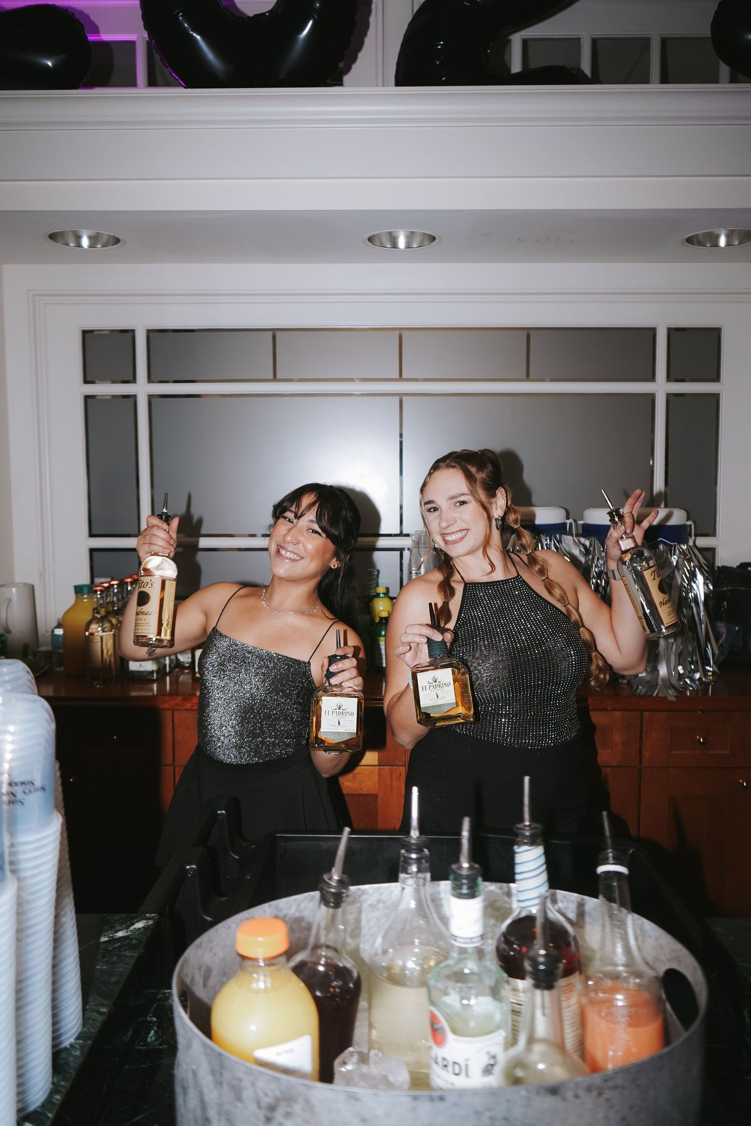 Two bartenders in matching sparkly outfits holding bottles behind a bar counter.