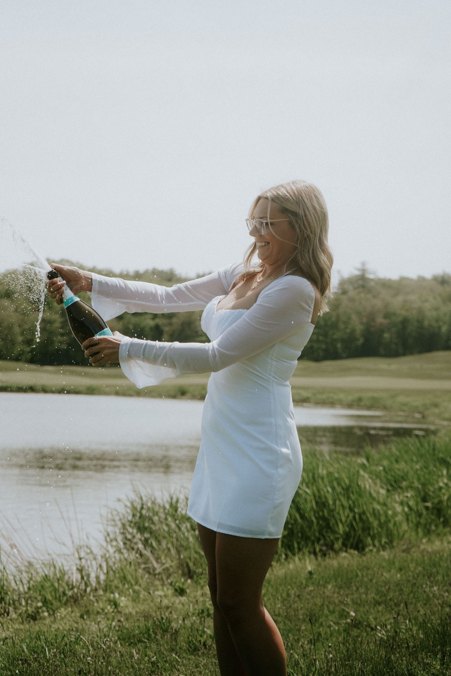 Woman in white dress celebrating with champagne by a serene lakeside.