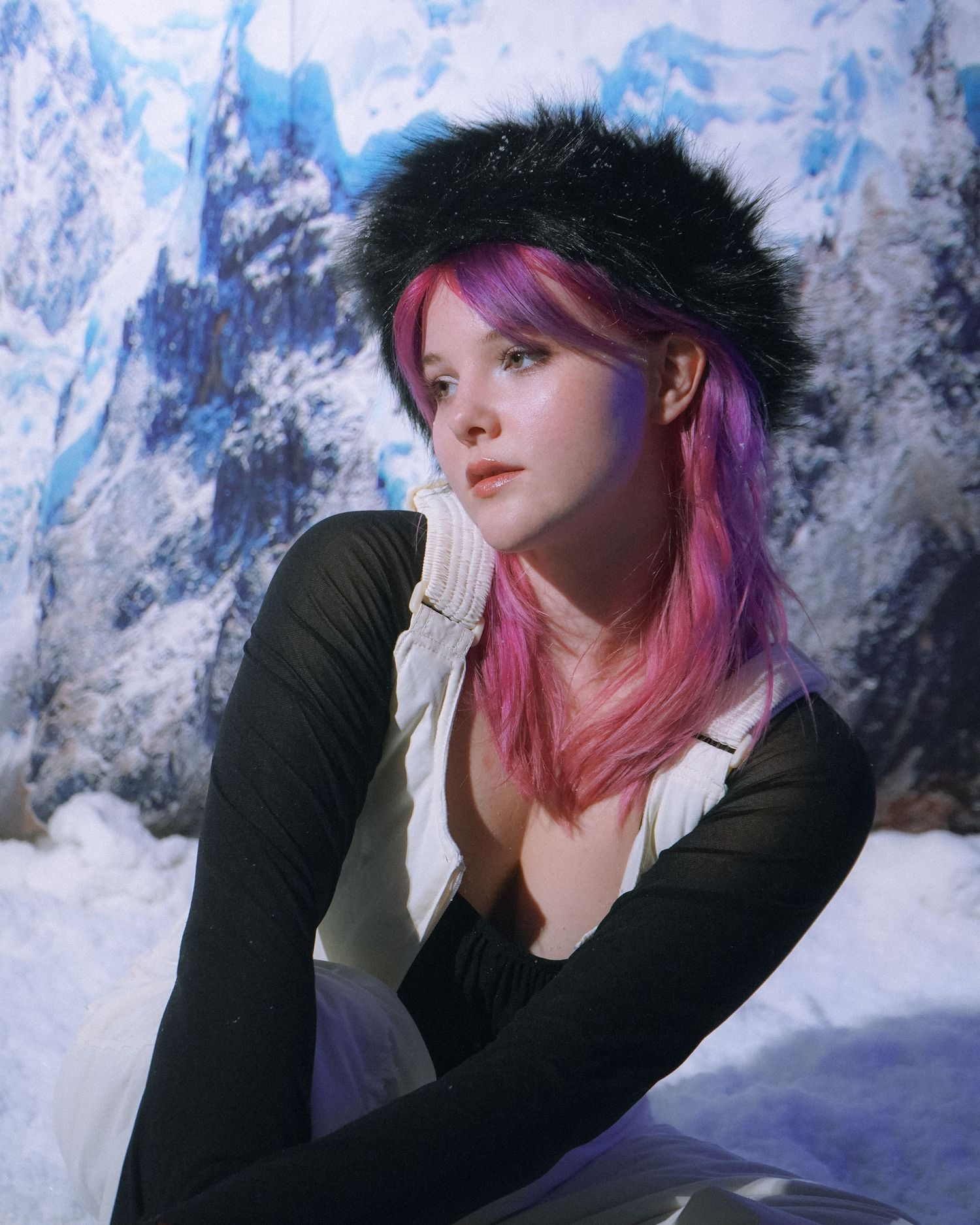 Winter fashion portrait featuring pink hair and fur hat against snowy mountain scene.