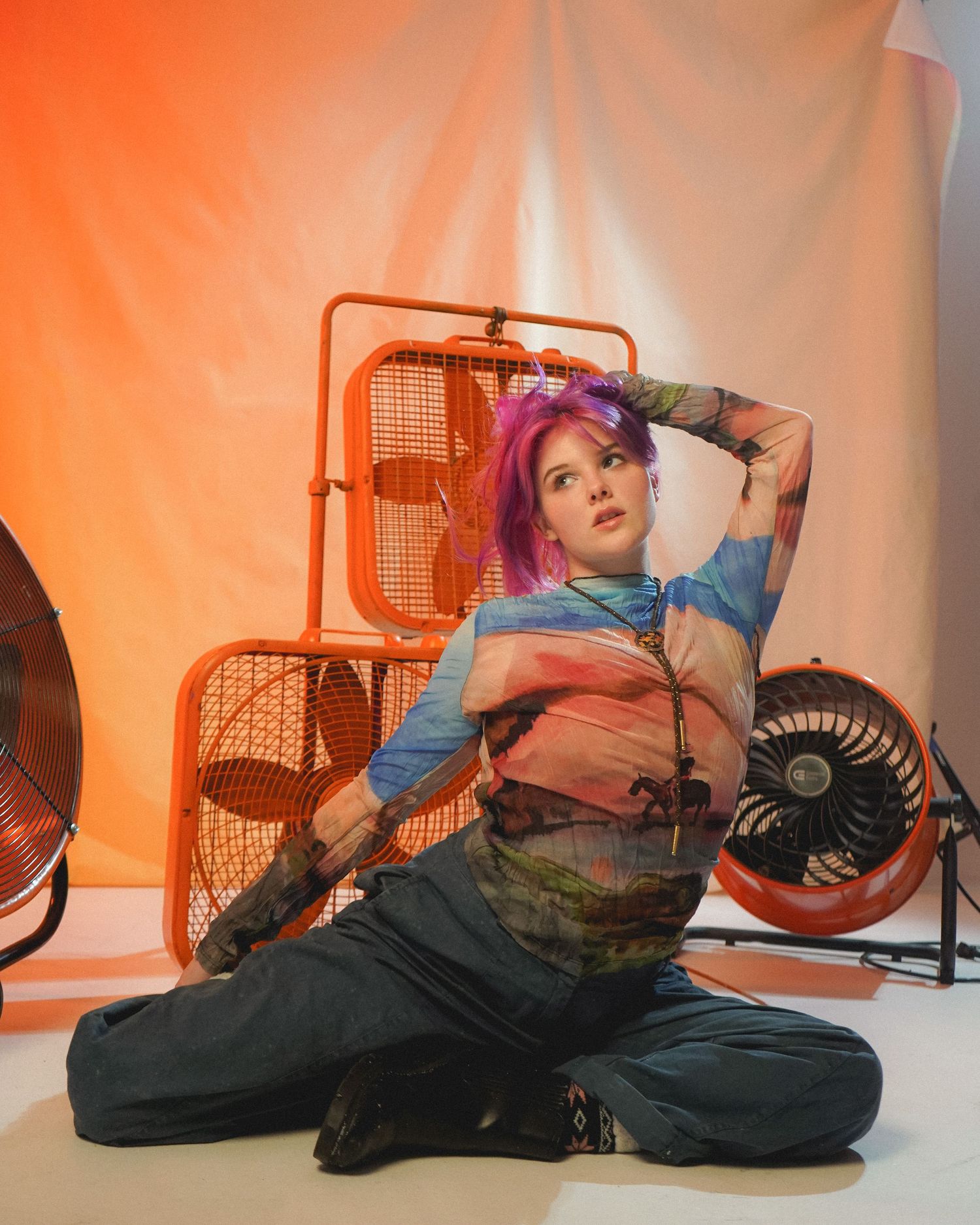 Model with pink hair strikes artistic pose among orange electric fans in studio setting.