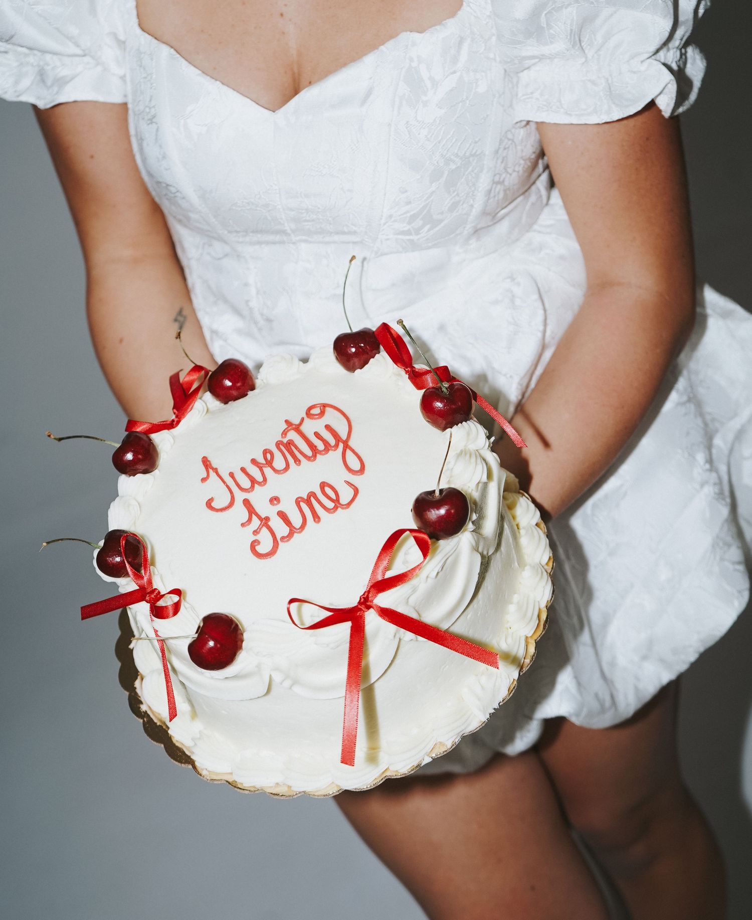 A white frosted cake decorated with fresh cherries and a red bow being held in a white dress.