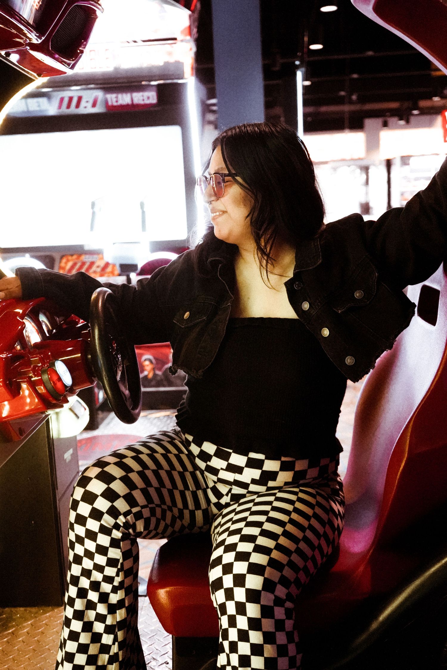 Someone plays an arcade racing game while wearing black jacket and checkered pants.