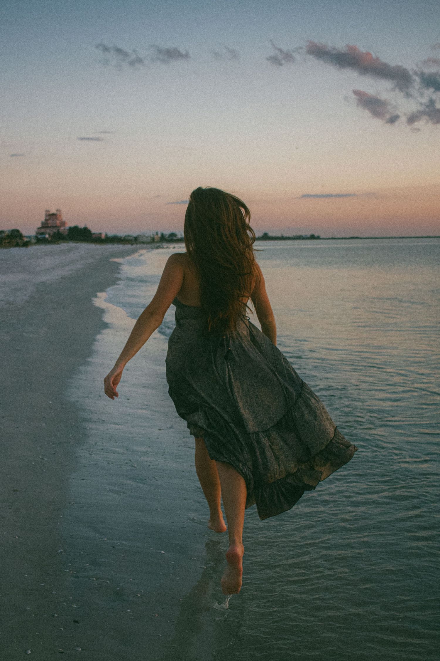 Woman in flowing dress walking along beach shoreline at sunset.