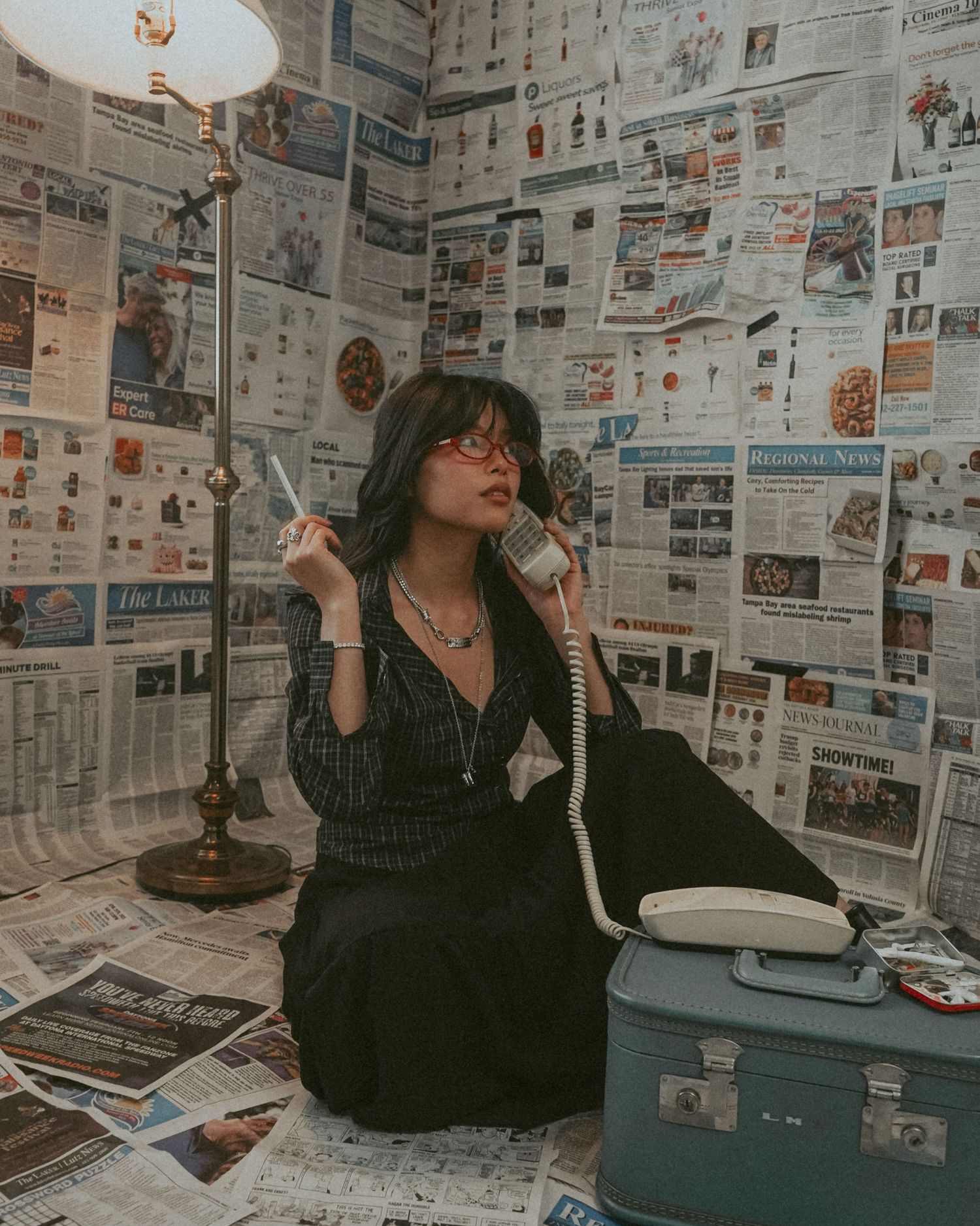 Vintage-styled photo with newspaper wall backdrop featuring corded telephone and desk lamp.