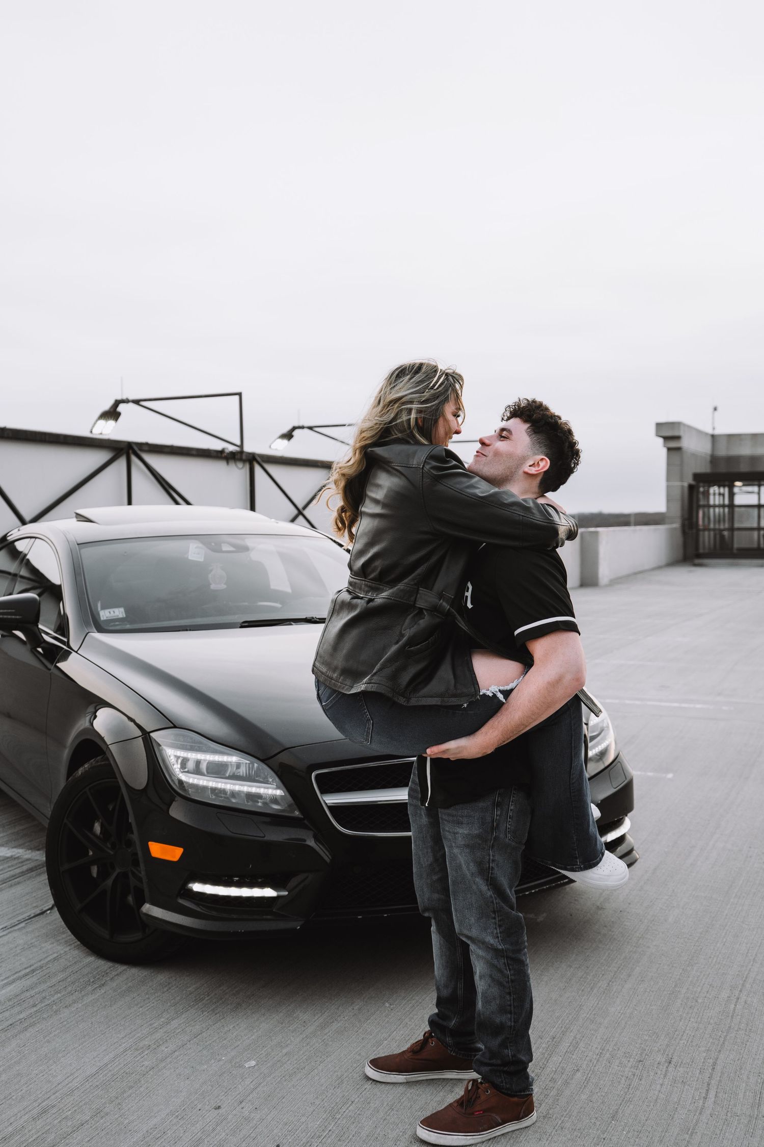 Romantic couple embracing in front of black Mercedes on rooftop parking garage.