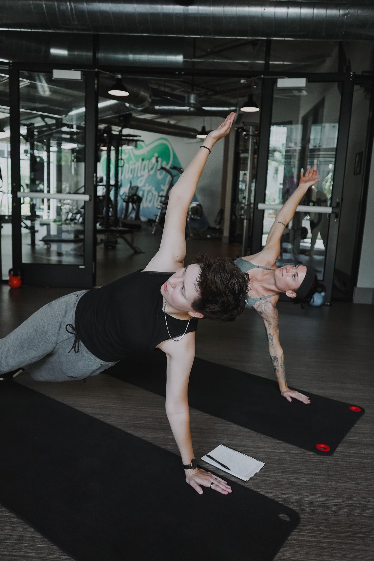 Two people performing side plank exercises on black exercise mats in gym.