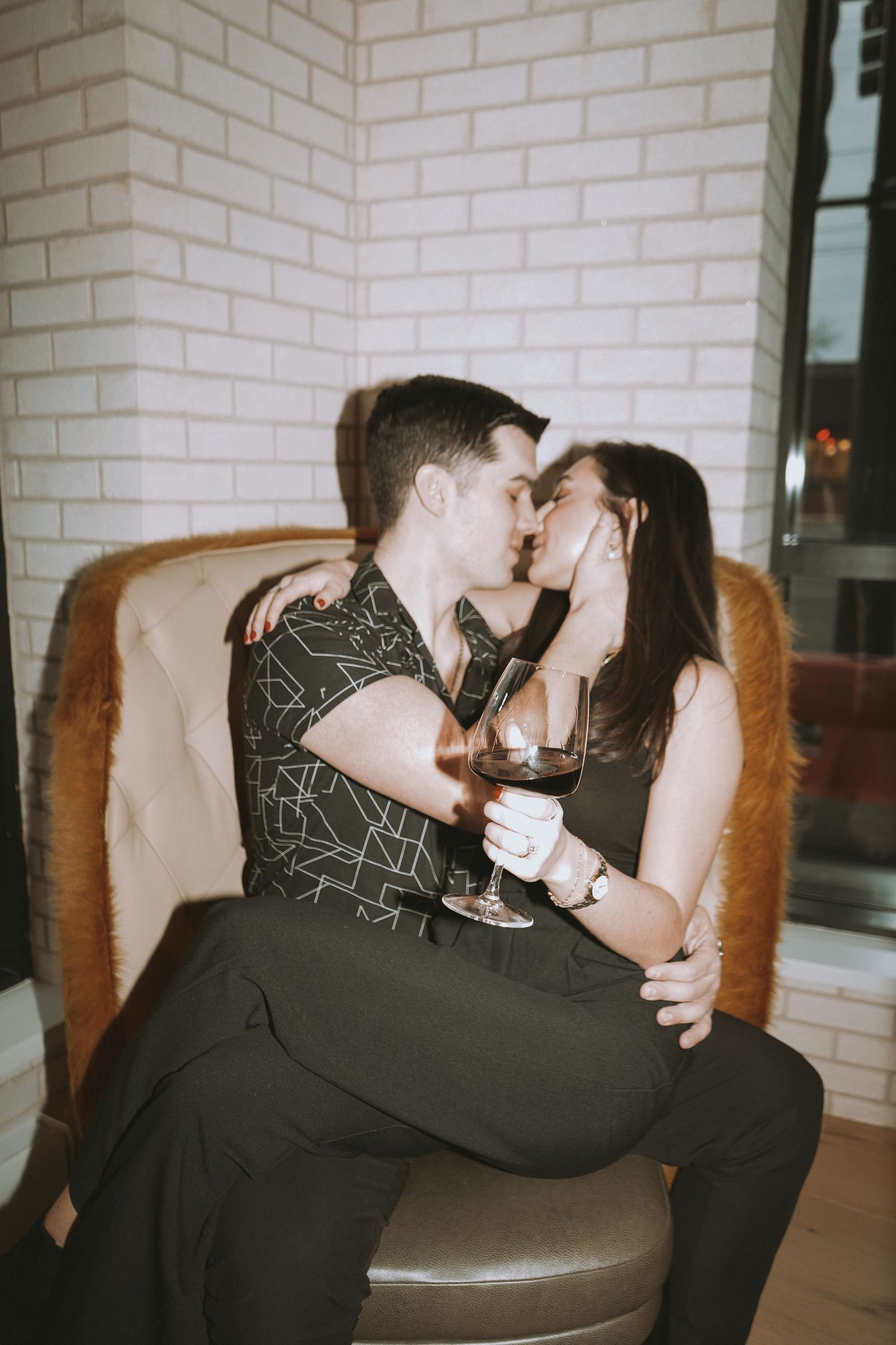 Intimate couple moment on brown leather chair against white brick wall.