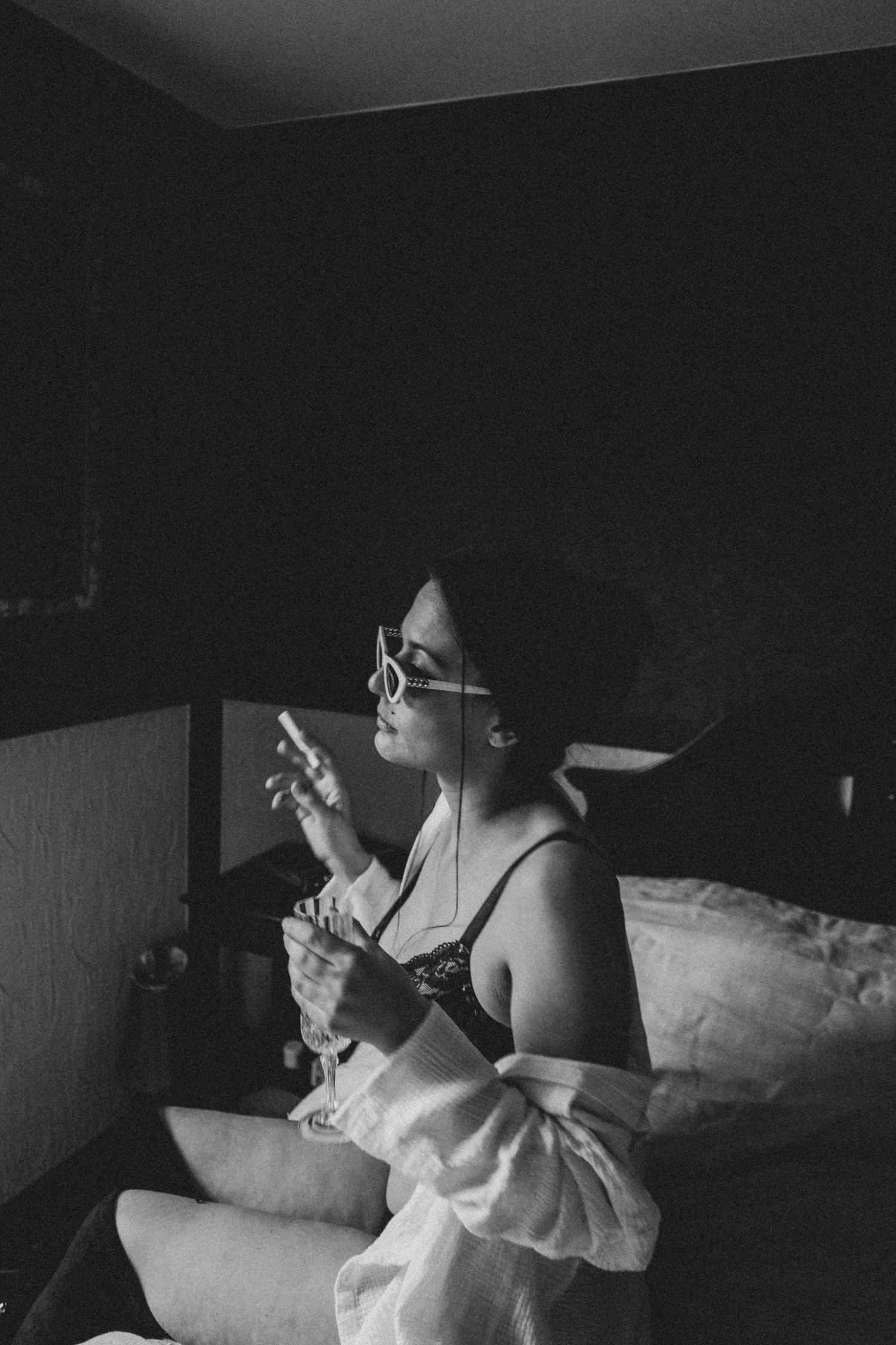 Black and white photo of someone sitting on bed wearing glasses and comfortable attire.