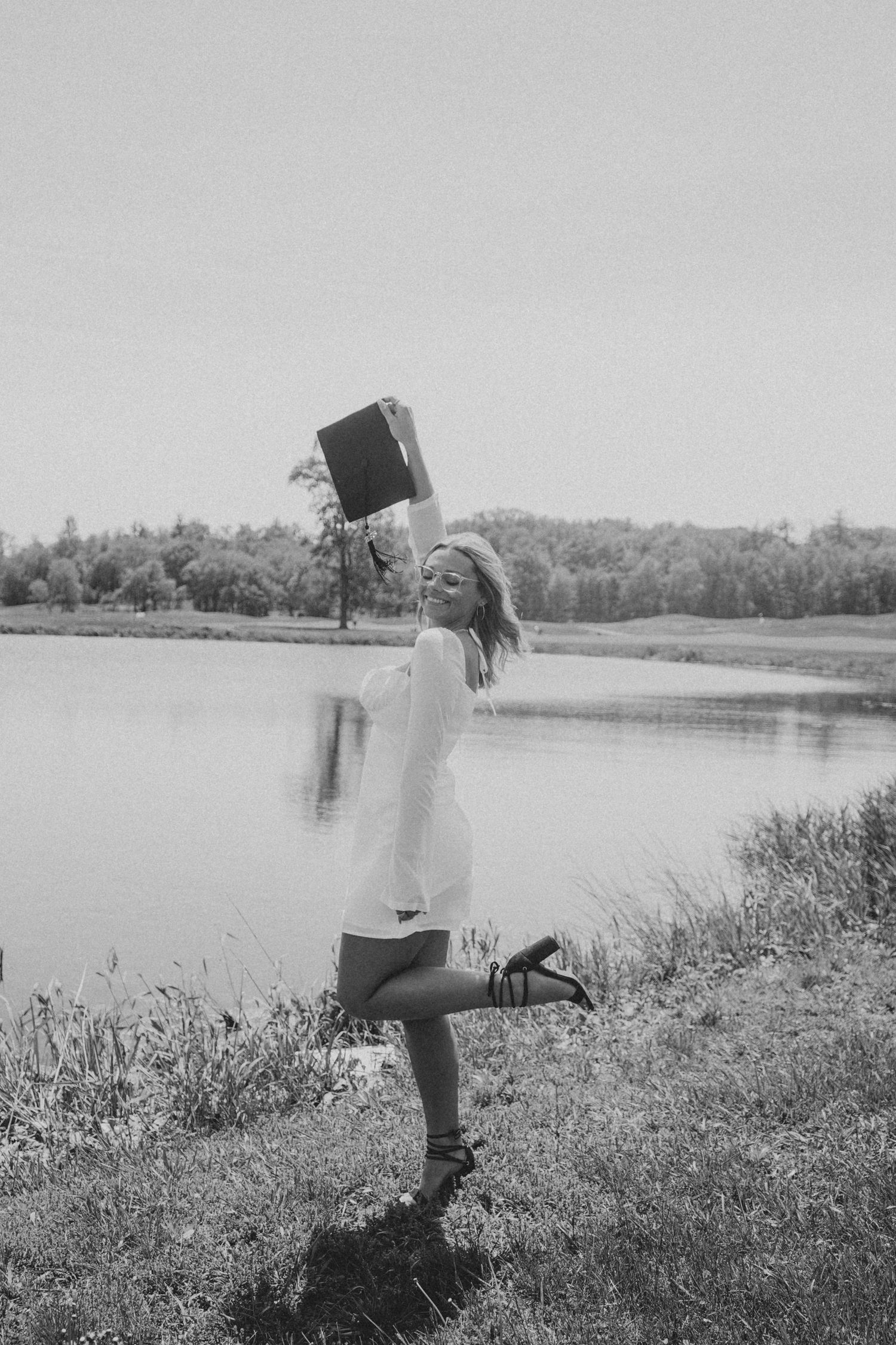 Woman in white dress joyfully holding a record player near a lake in black and white.