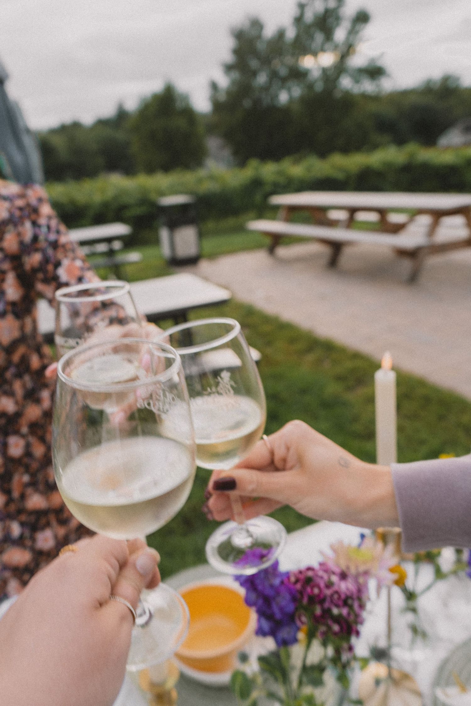 Two hands toasting with wine glasses over an outdoor table decorated with colorful flowers.
