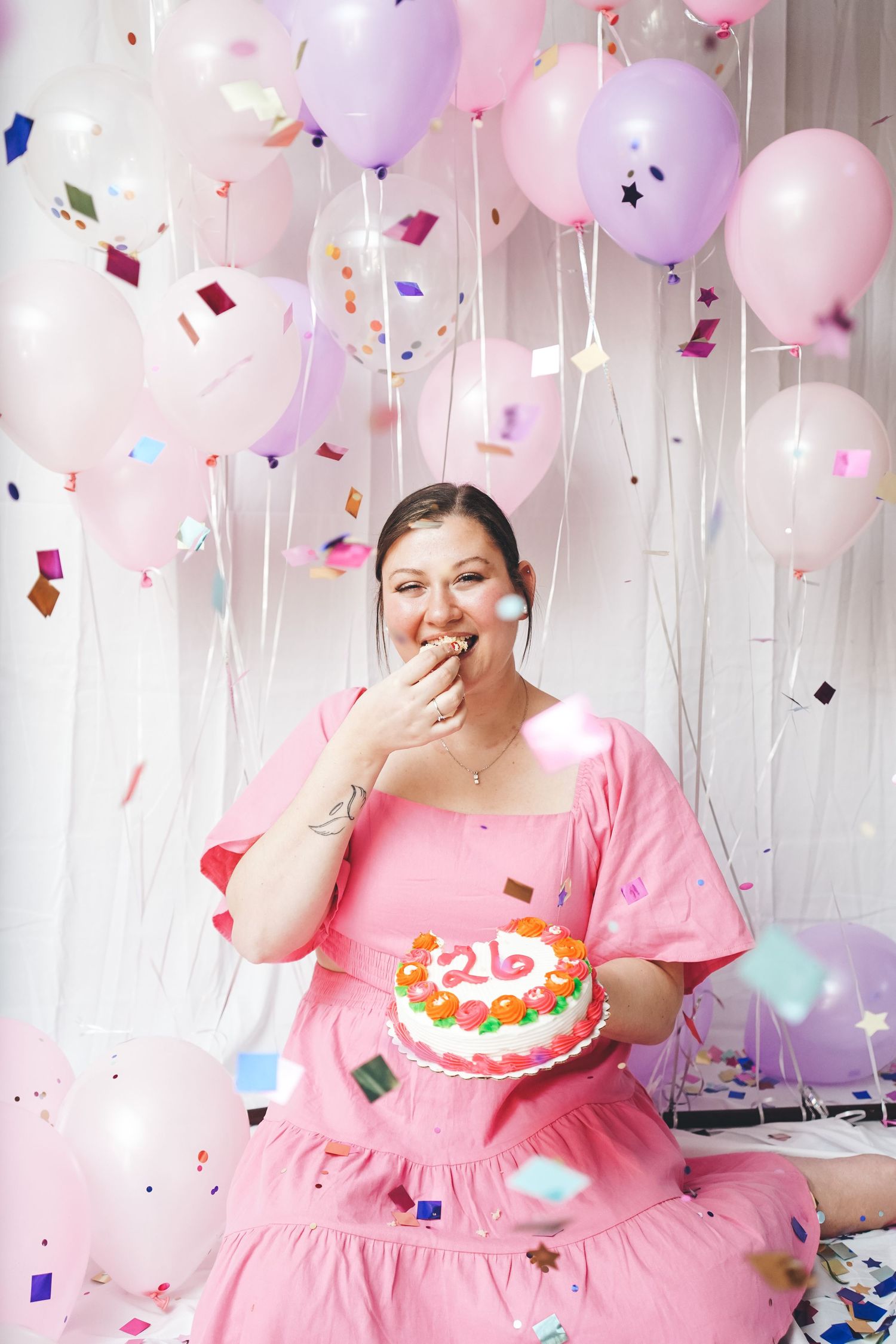 Woman in pink dress enjoys birthday celebration with cake amid pink and purple balloons.