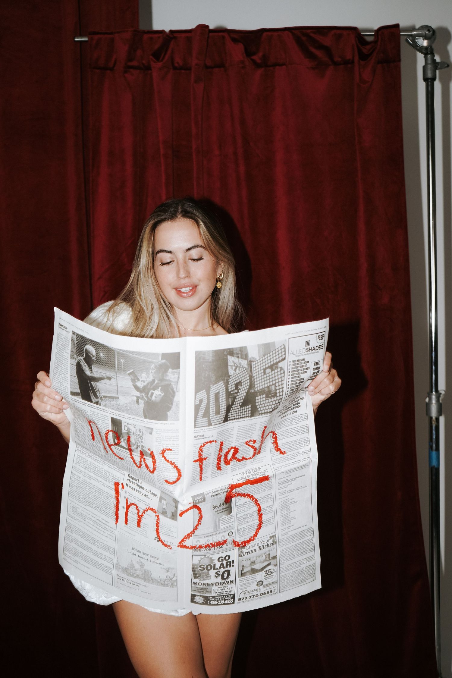Casual portrait reading newspaper against red curtain backdrop in studio setting.