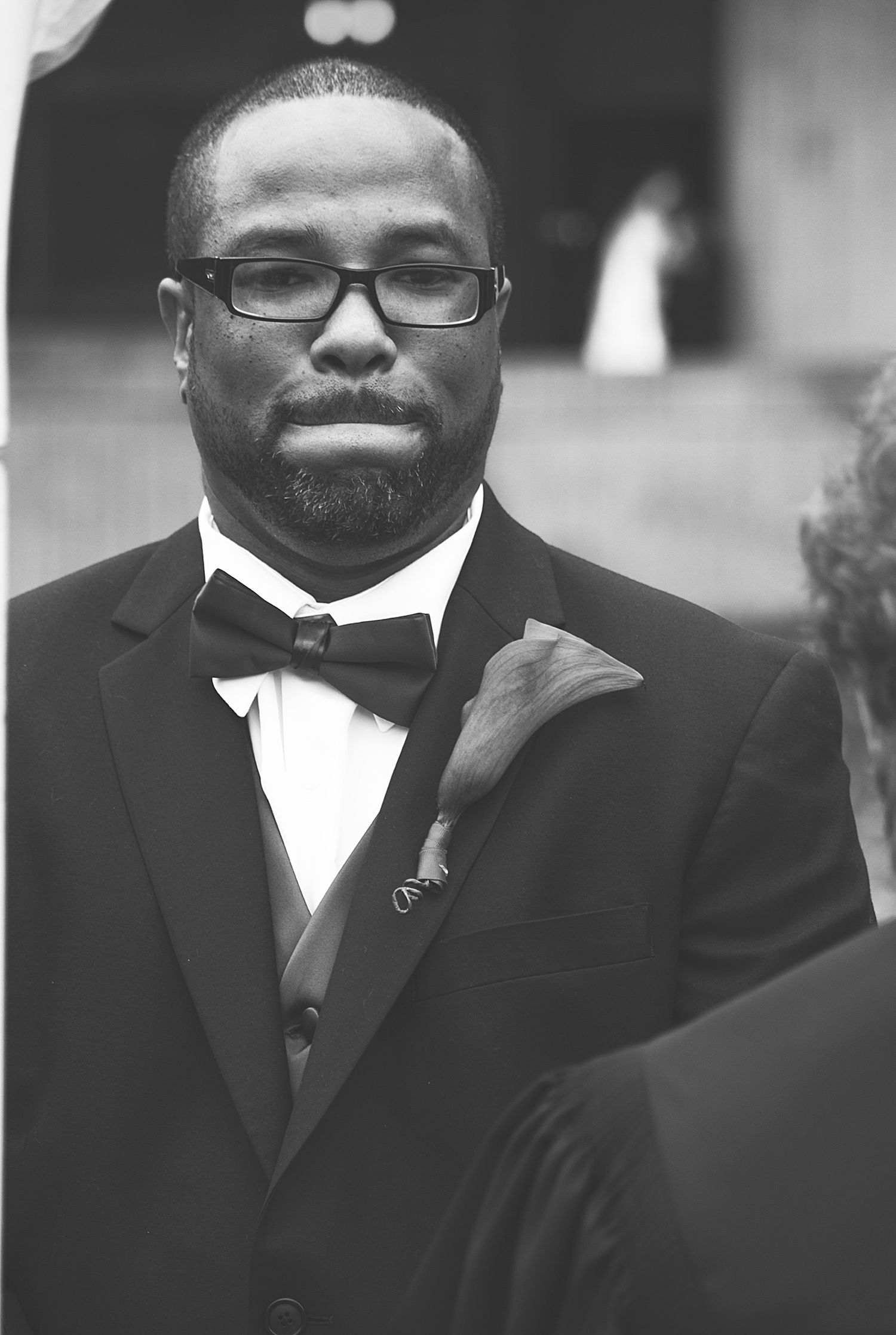 Formal portrait of someone wearing a black tuxedo with bow tie and glasses in black and white.