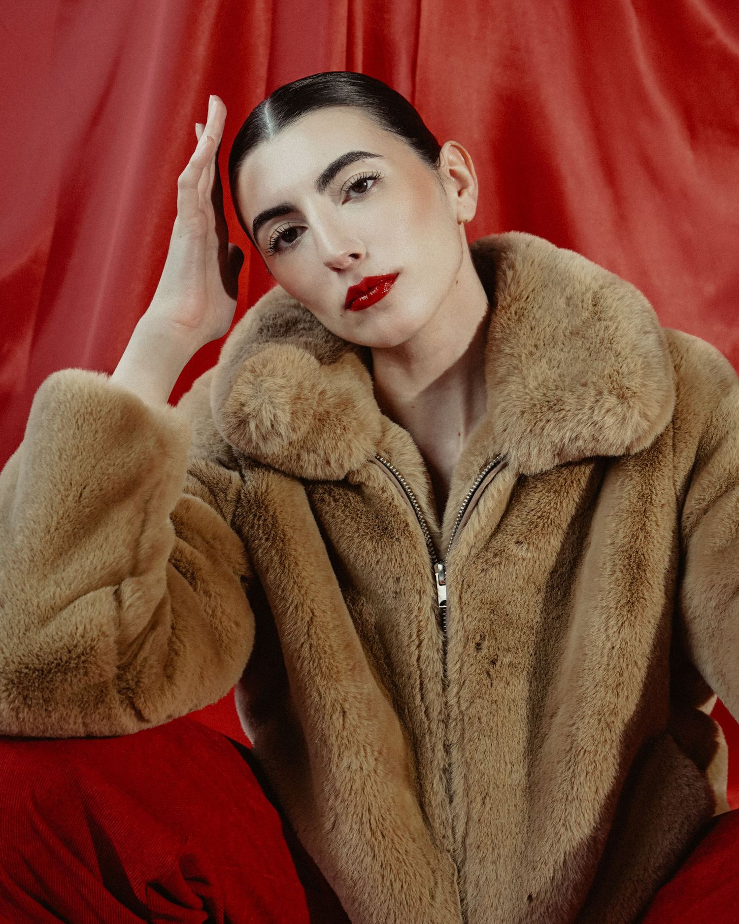 Fashion model in tan fur coat poses against red backdrop wearing bold red lipstick.