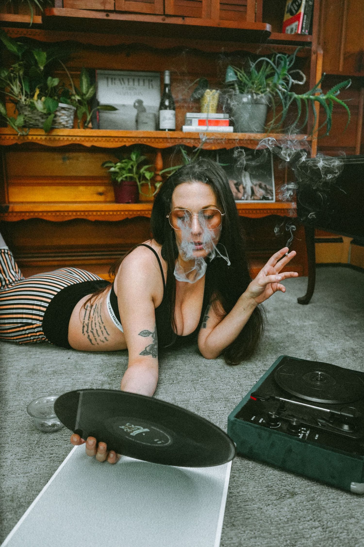 Person poses with vinyl record on carpet near wooden shelves with houseplants.