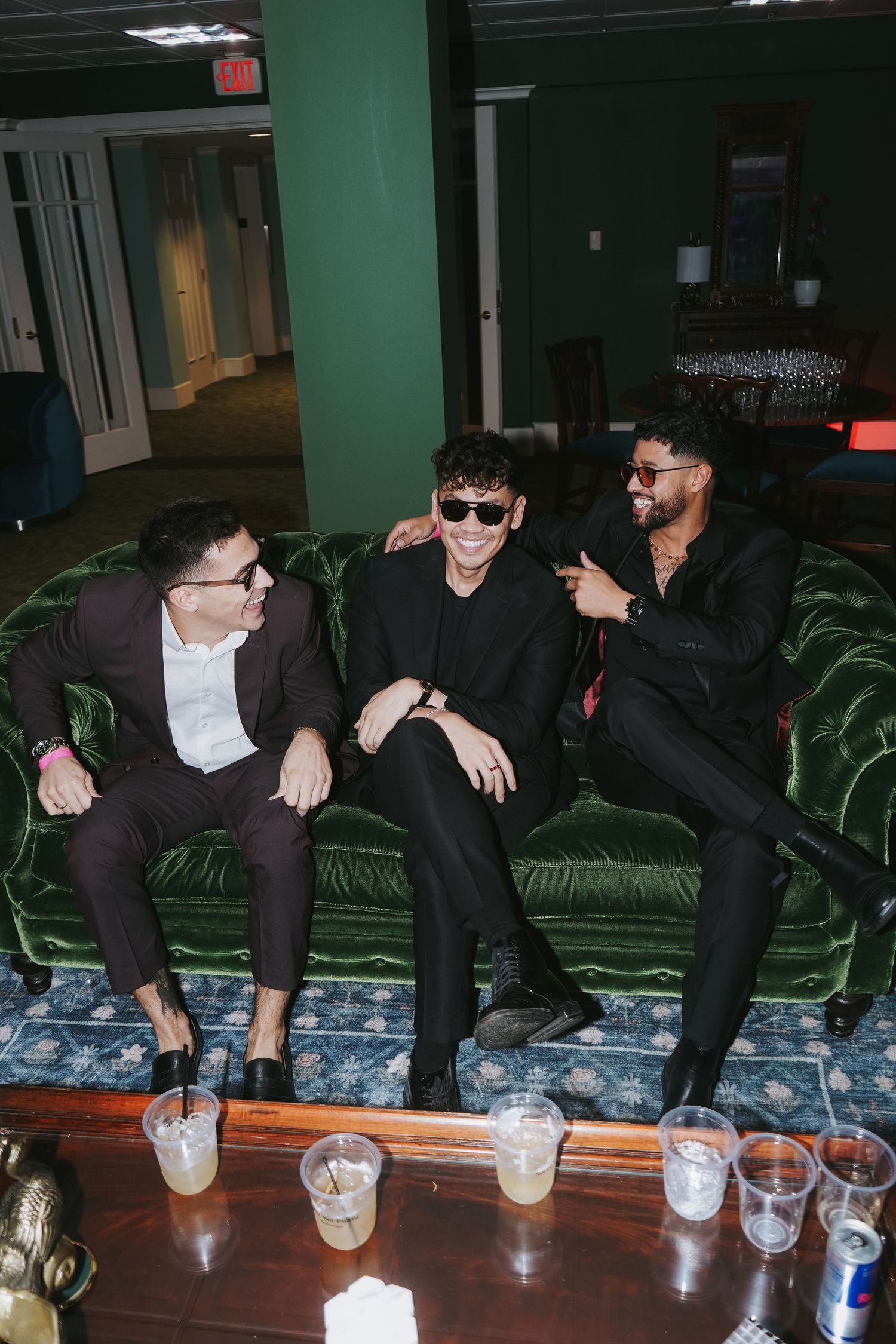 Three people socializing on a green velvet couch with drinks on the coffee table.