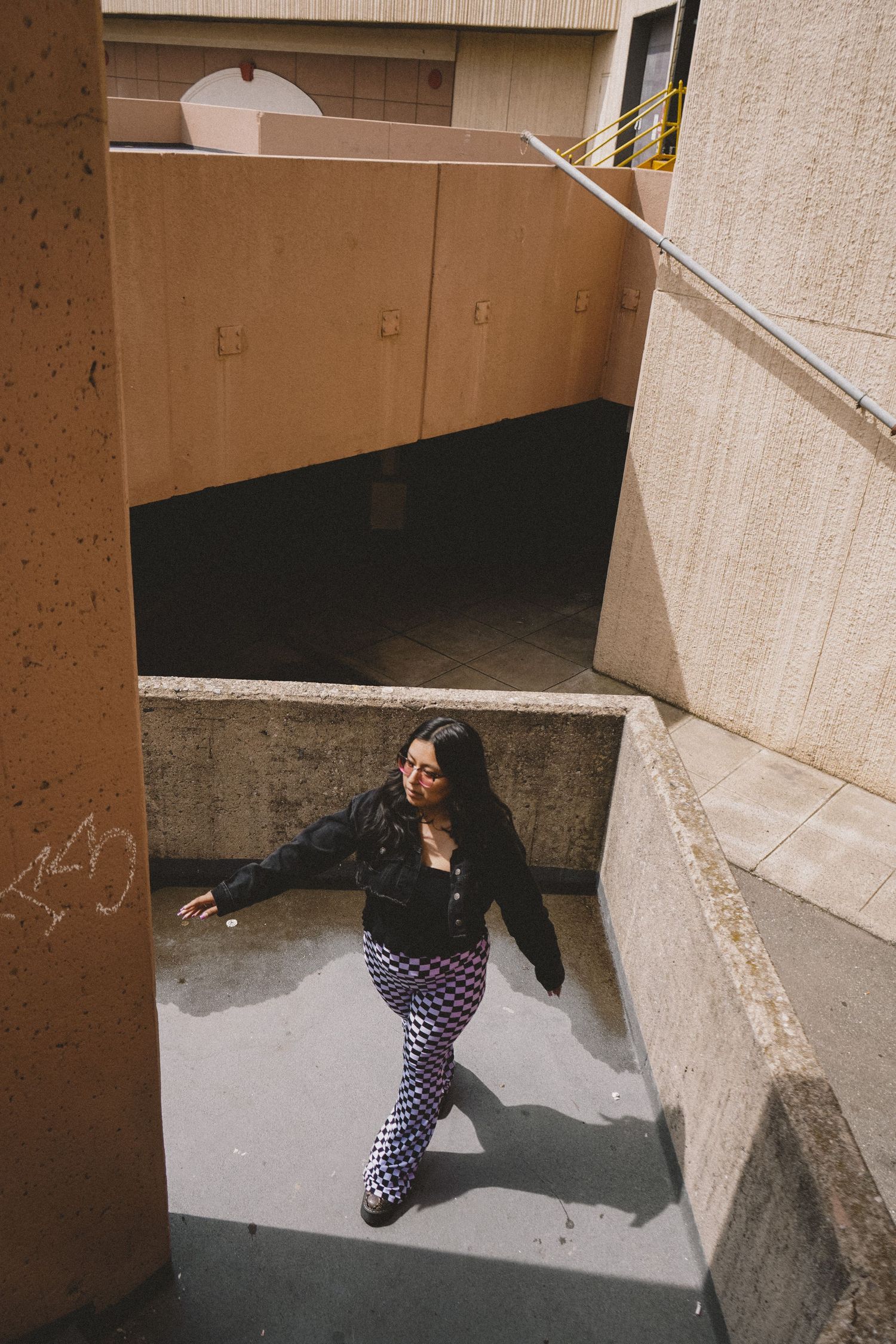 Person in checkered pants and black jacket poses in concrete stairwell with dramatic lighting.