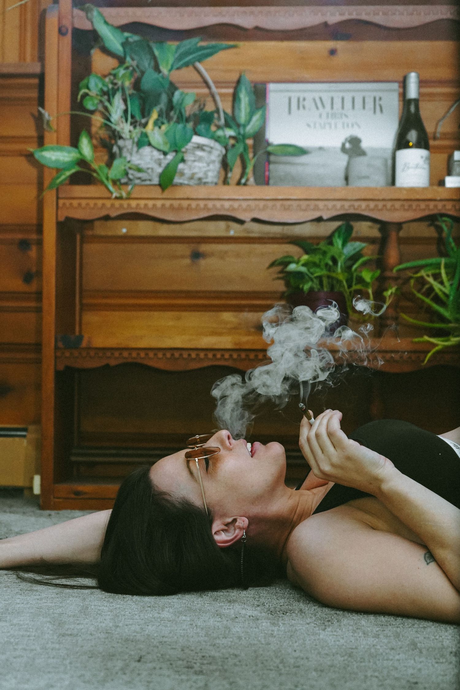 Person exhaling smoke while lying on floor with vintage furniture and plants in background.
