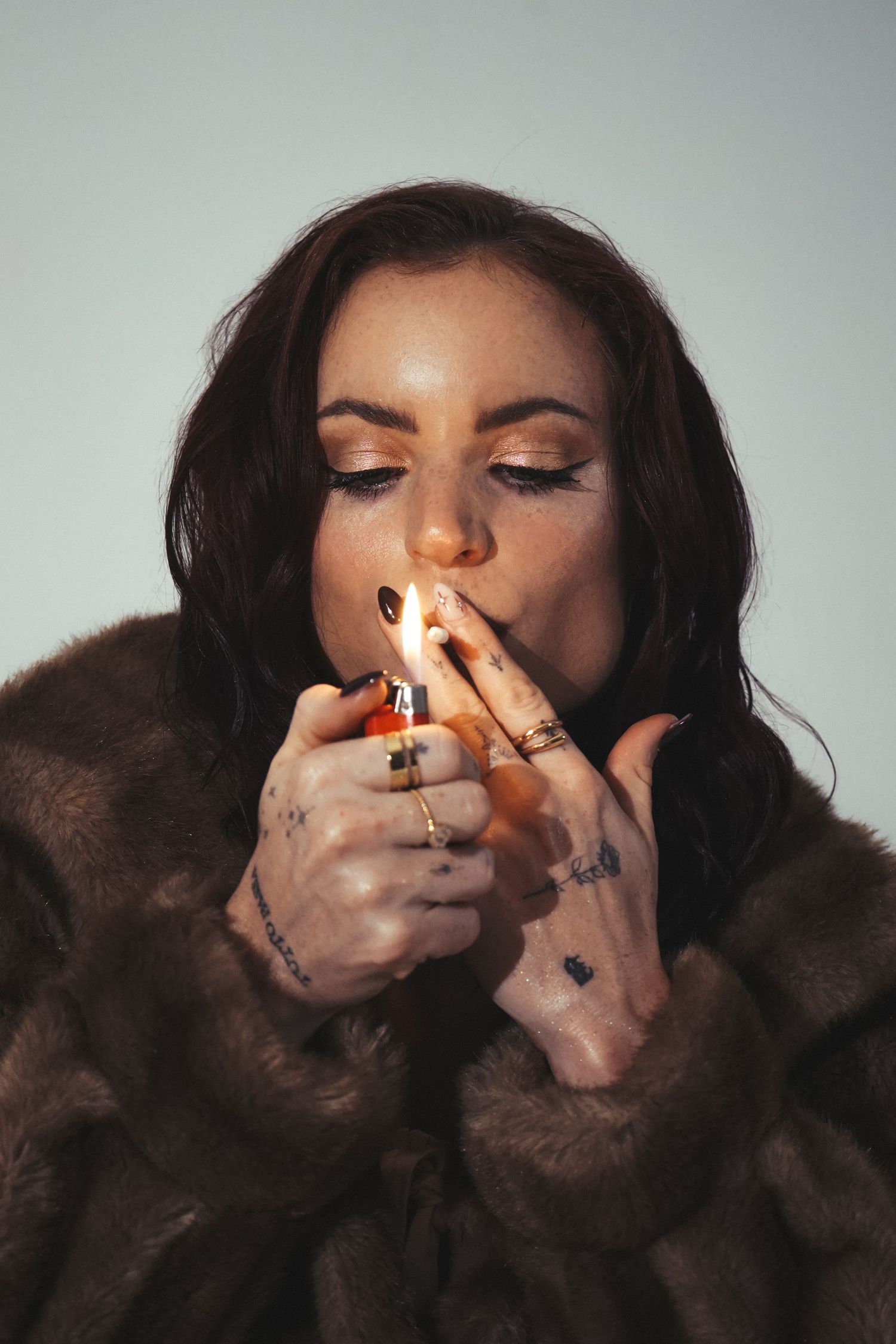 Person in brown fur coat lighting a cigarette with eyes closed and wearing glamorous makeup.