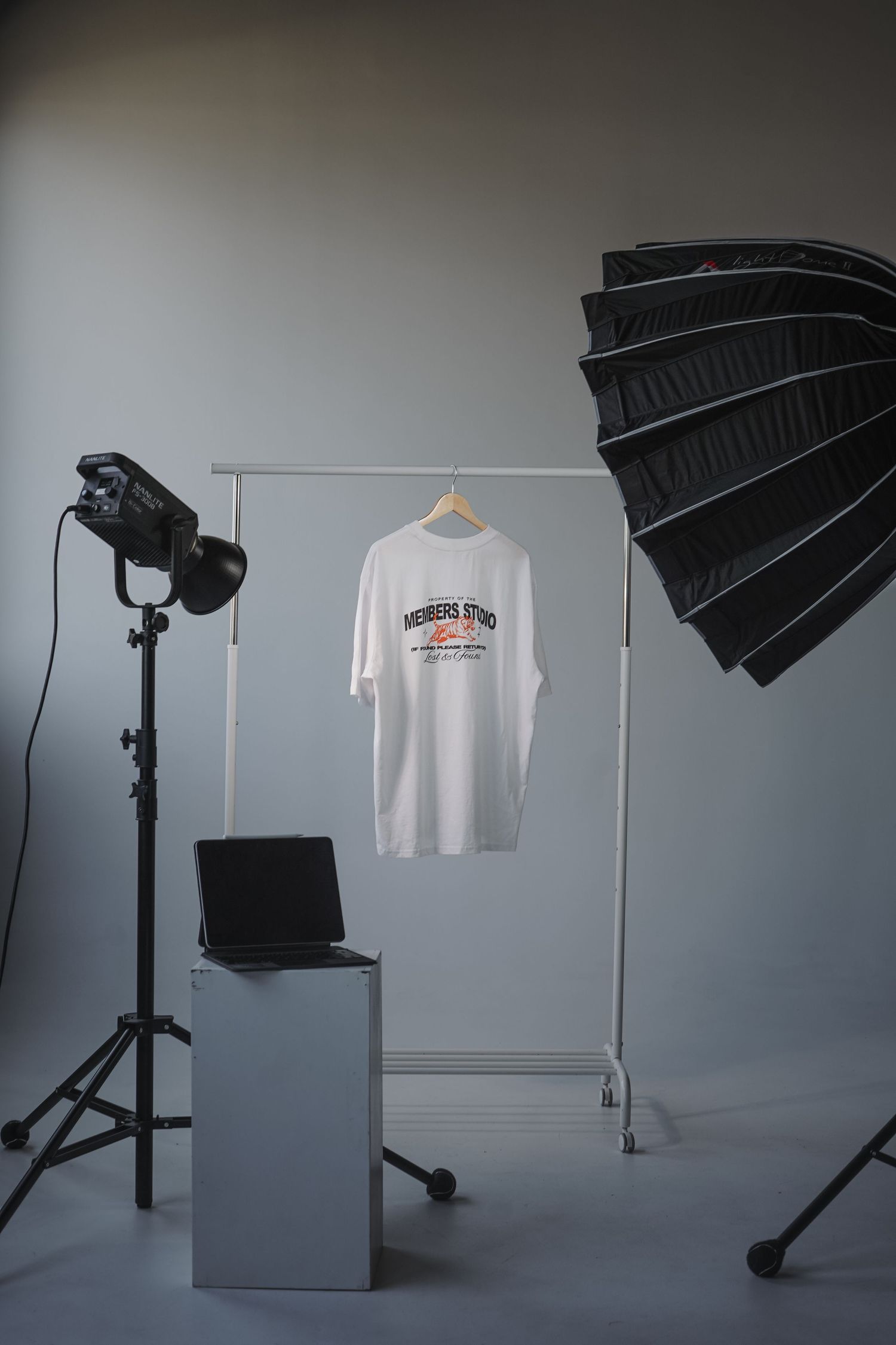 Photography studio setup with lighting equipment, laptop, and white t-shirt hanging on clothing rack against gray backdrop.