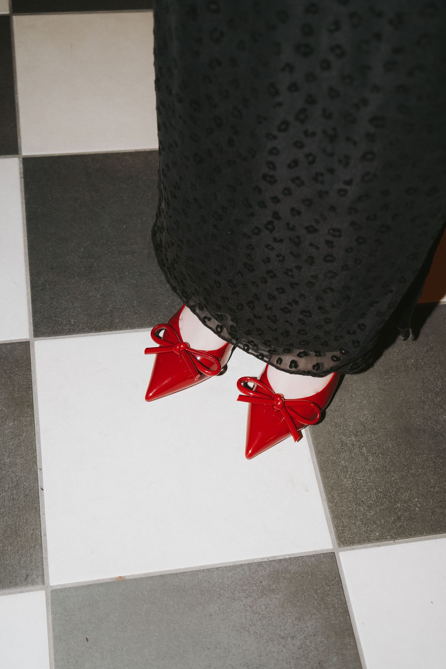 Red pointed high heels peek out from under black lace pants on a checkered floor.