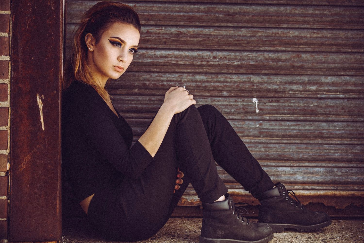 Fashion portrait of woman in all black outfit sitting against rustic wooden wall.