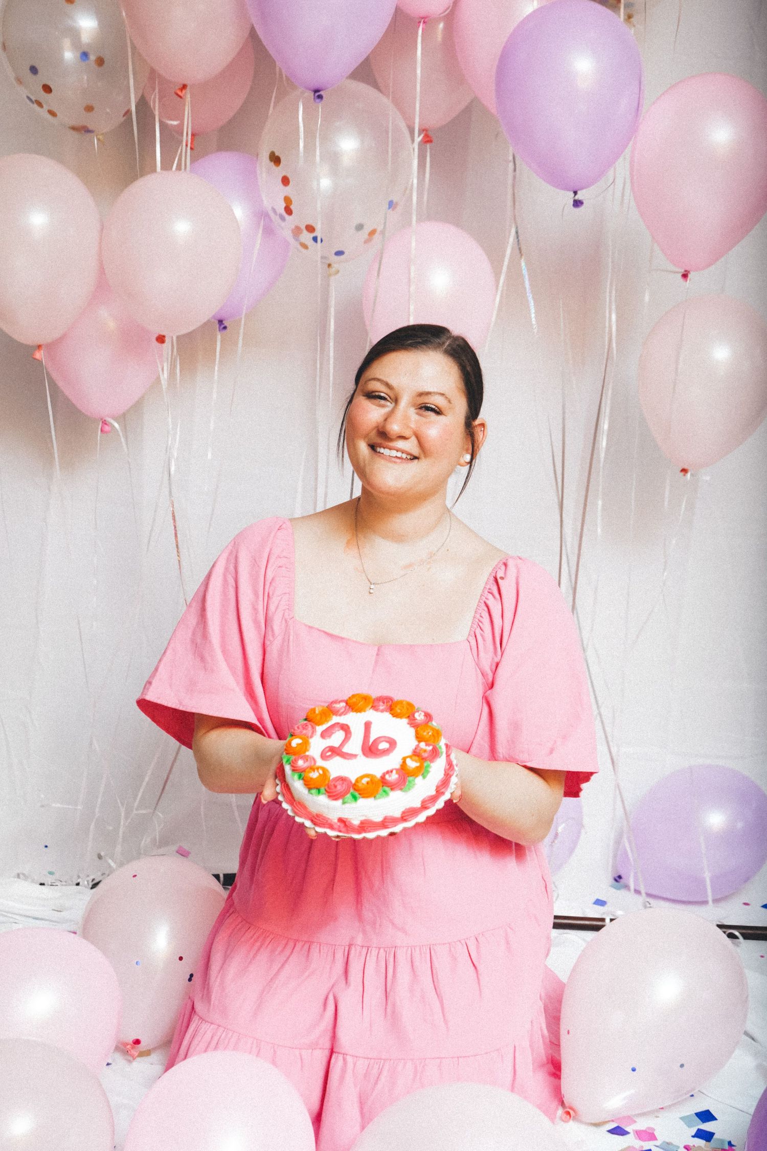 Woman in pink dress holds birthday cake while surrounded by pastel balloons at party.