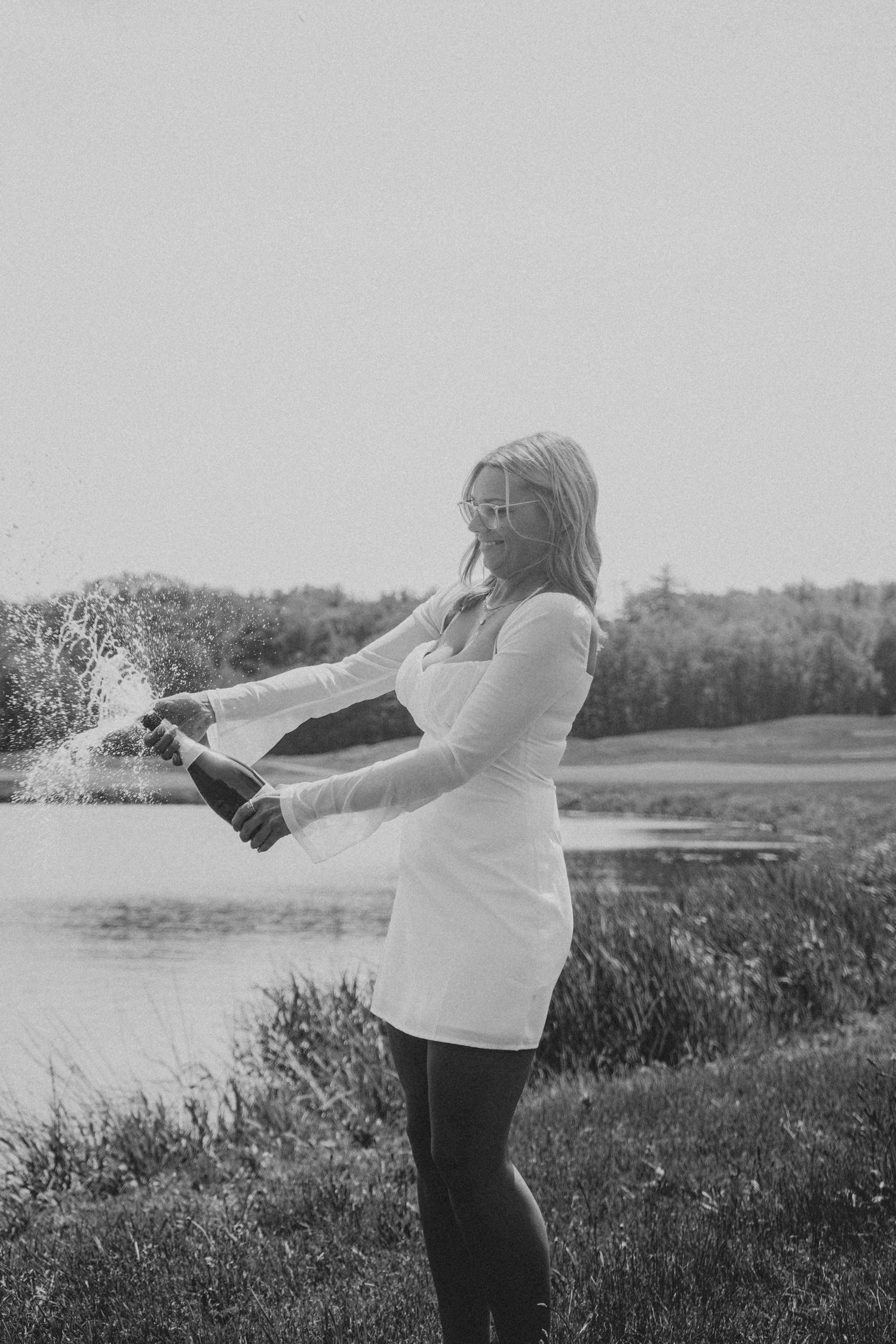 Woman spraying champagne outdoors during a celebratory moment by the water.