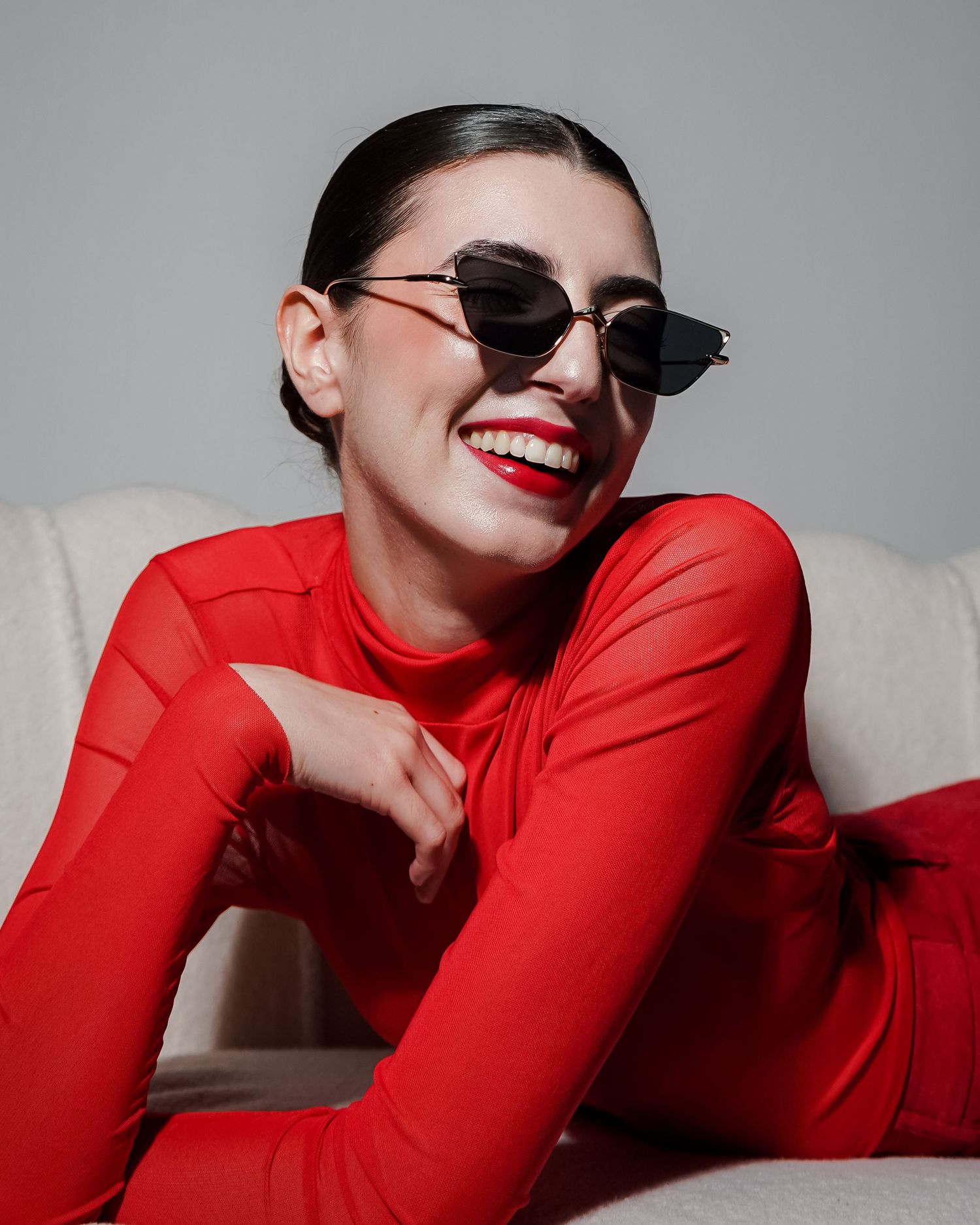 Fashion model in red outfit and sunglasses poses playfully on couch.