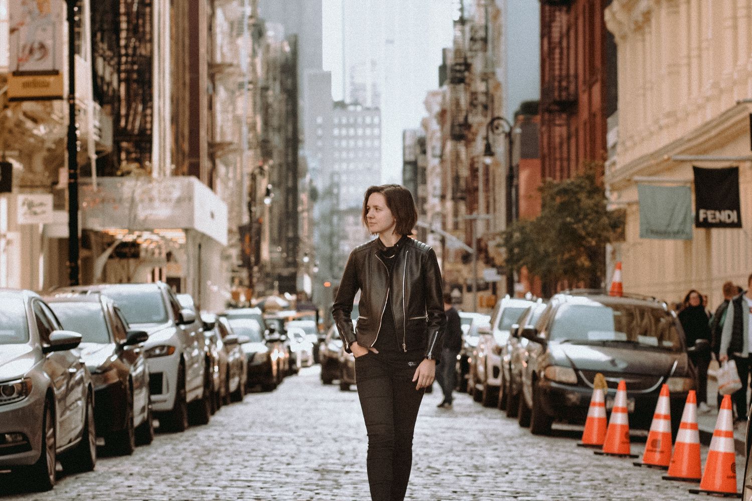 Person walking down cobblestone street lined with parked cars in urban setting.