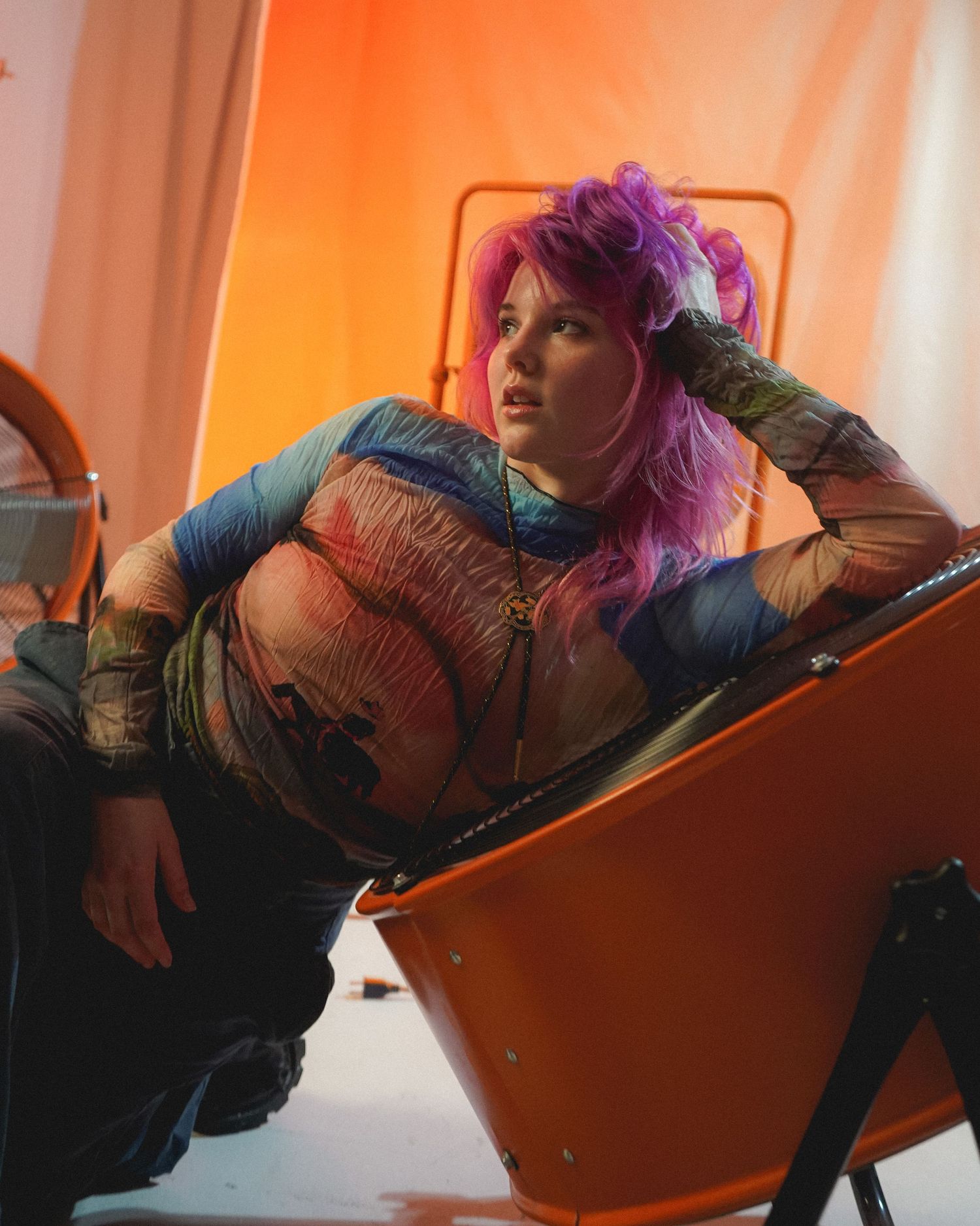 Model with vibrant pink hair reclines in orange chair wearing colorful streetwear.