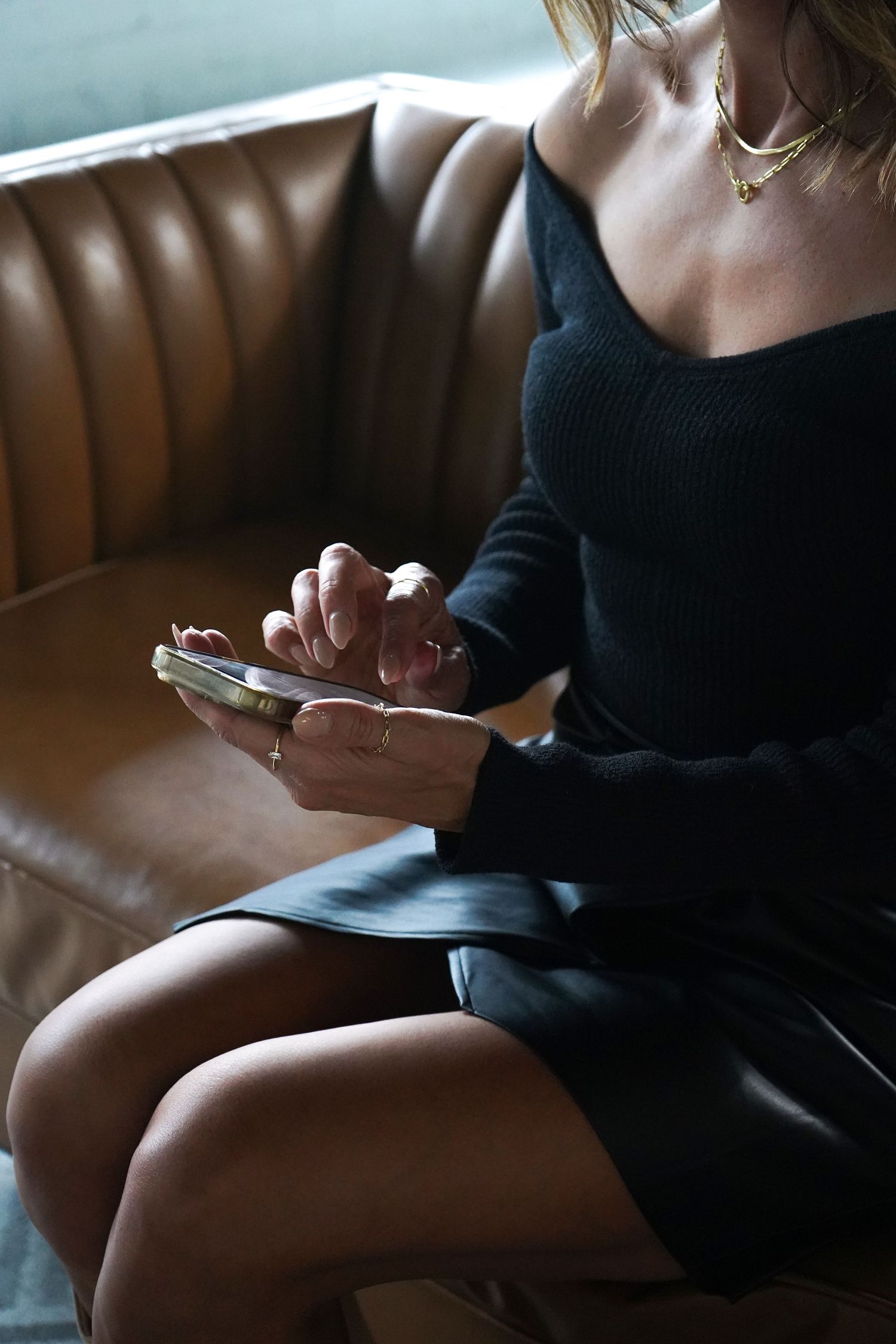 Woman in black dress sitting on leather couch using smartphone.