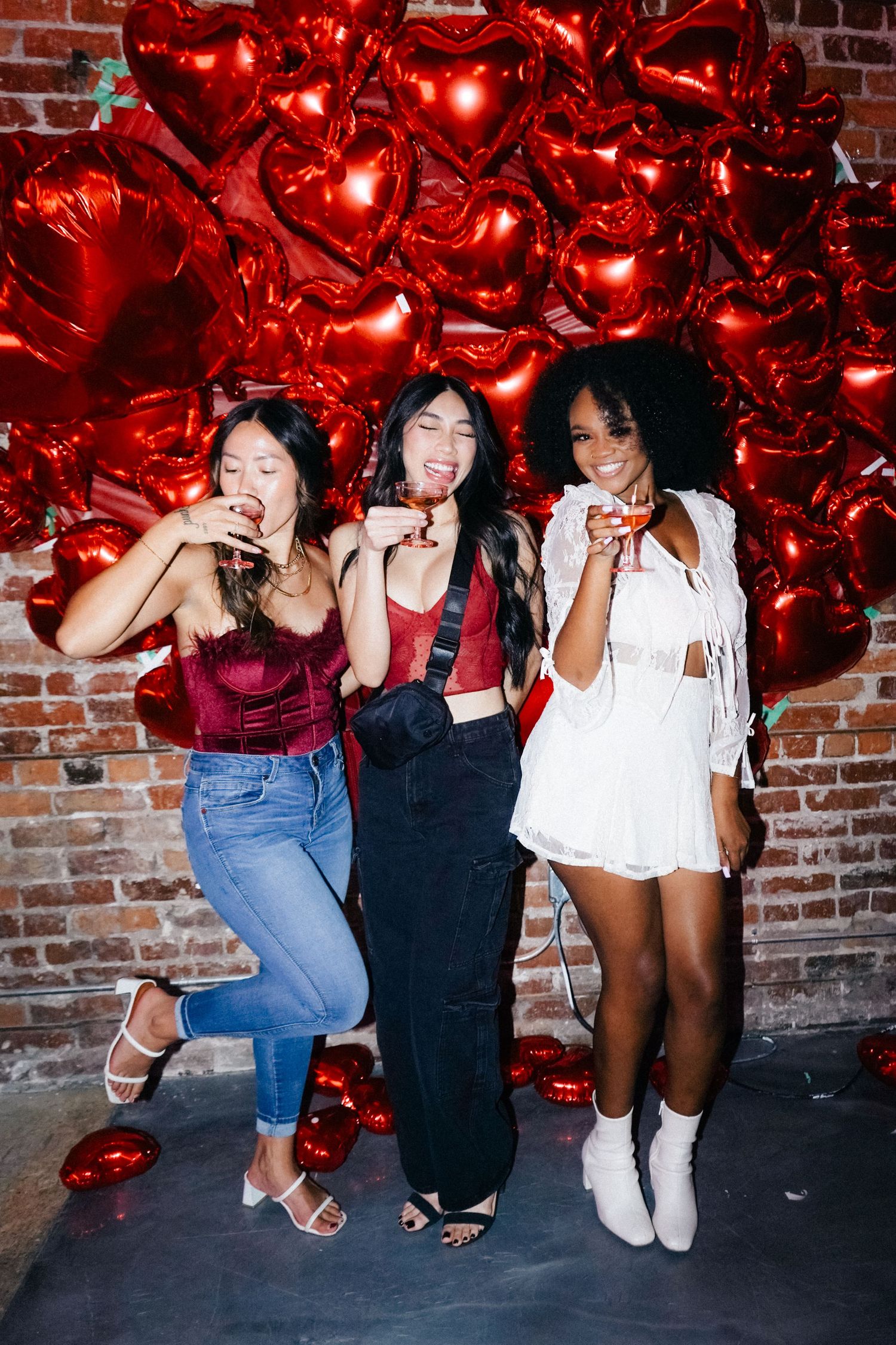 Three friends posing and laughing against red heart balloon backdrop at party.
