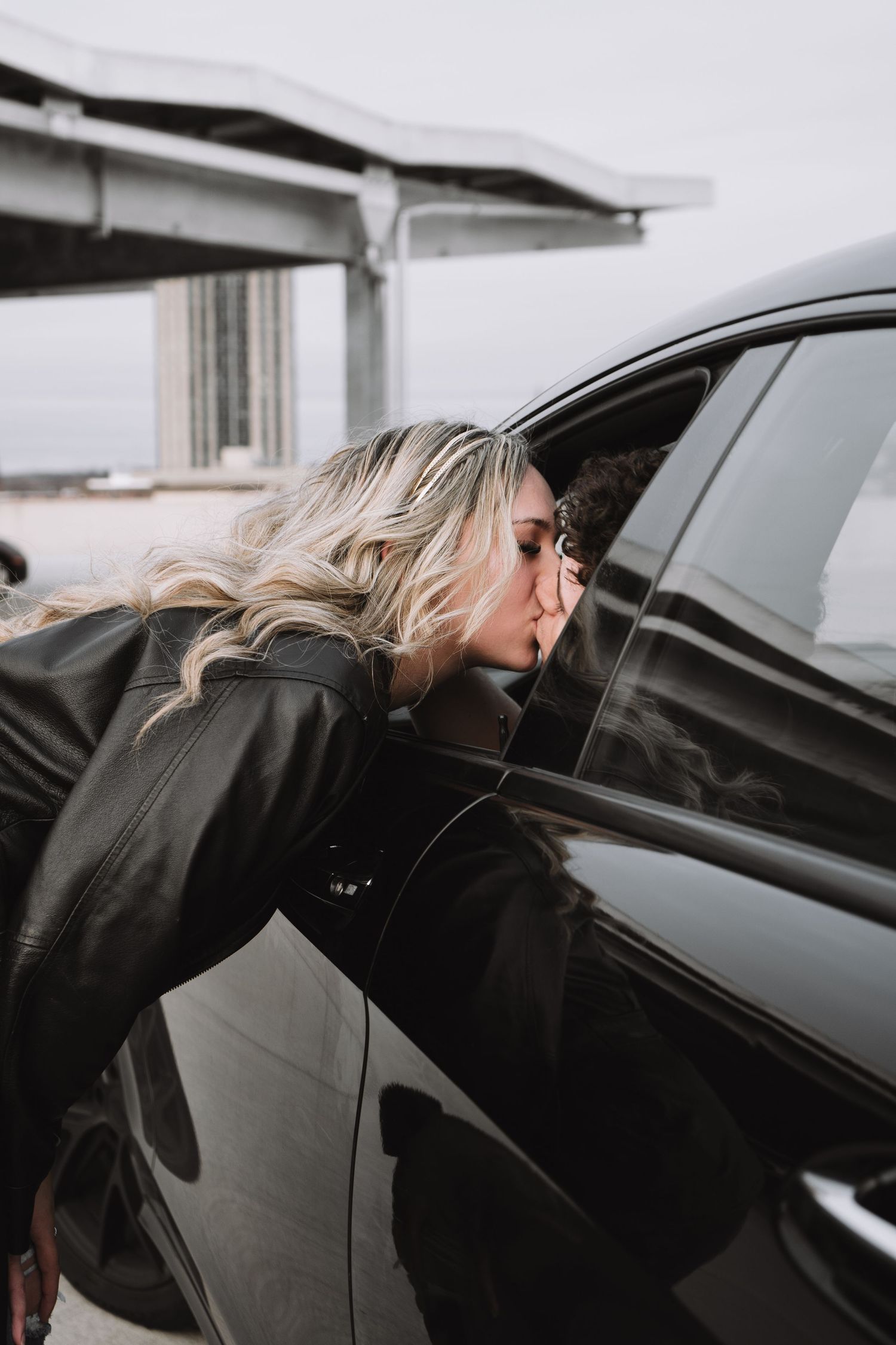 Blonde figure in black jacket leaning into dark car window on overcast day.