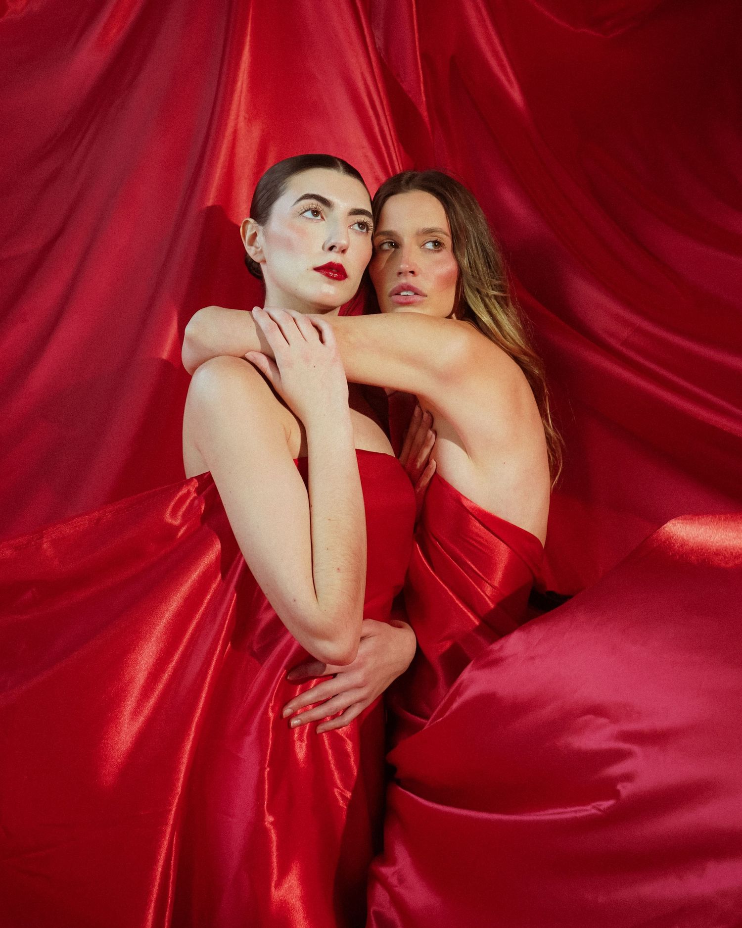 Two models pose dramatically against flowing red satin fabric in an artistic fashion photoshoot.
