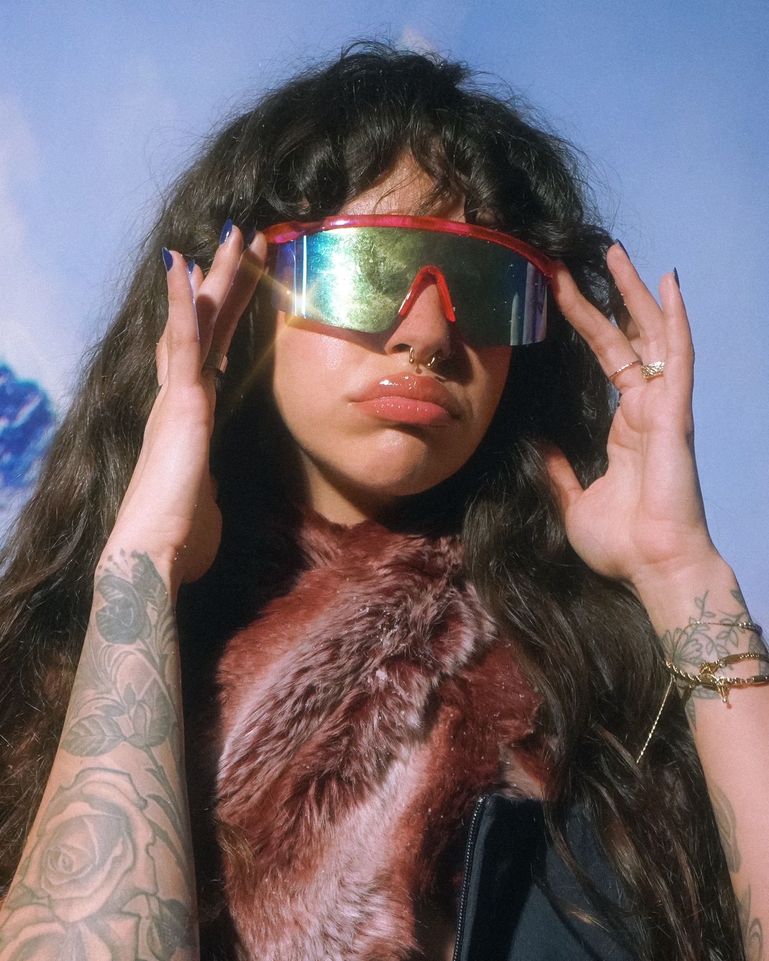 Model with tattoos and long hair wears iridescent sunglasses against snowy mountain backdrop.