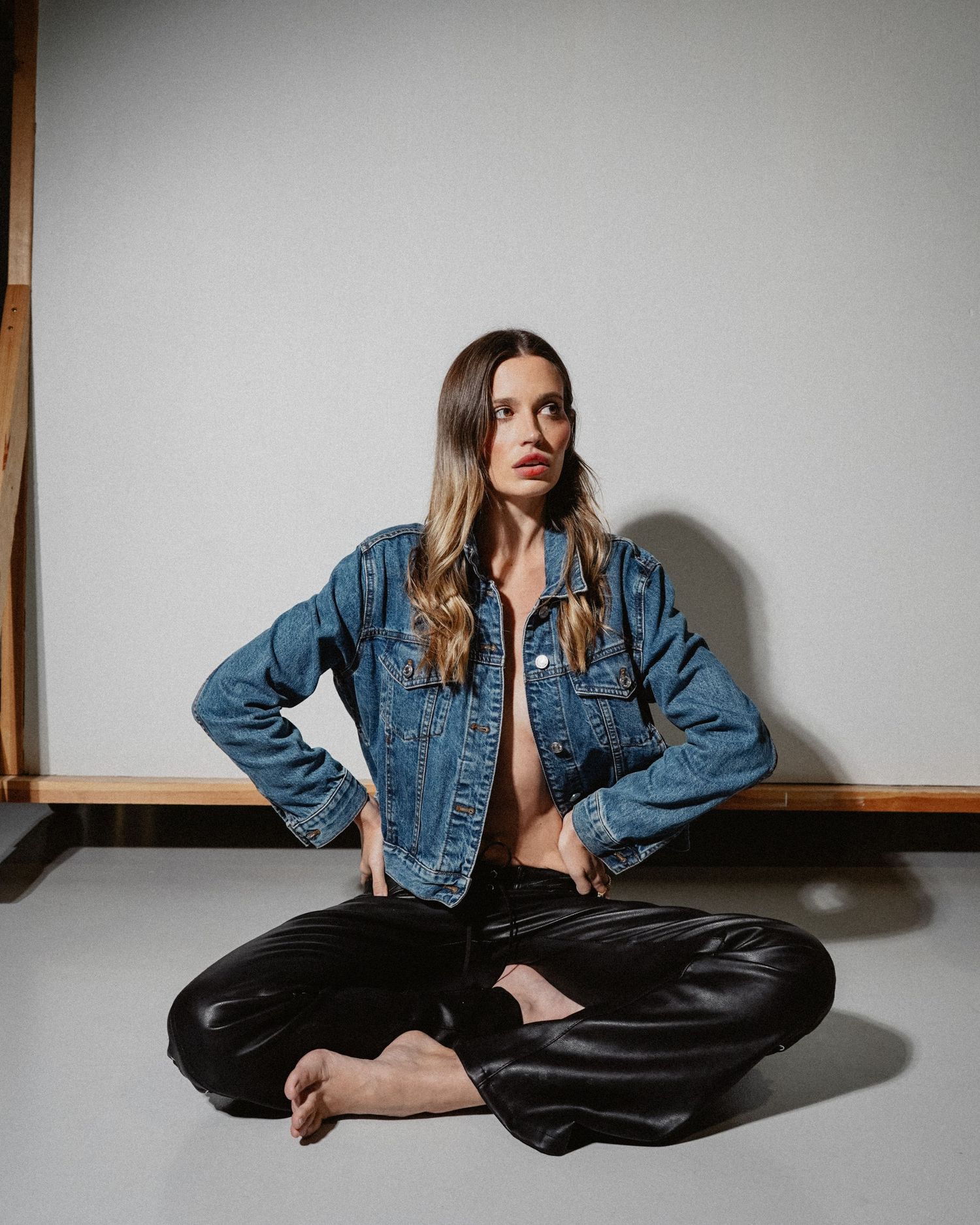 Fashion model poses in denim jacket and leather pants against white backdrop with moody lighting.