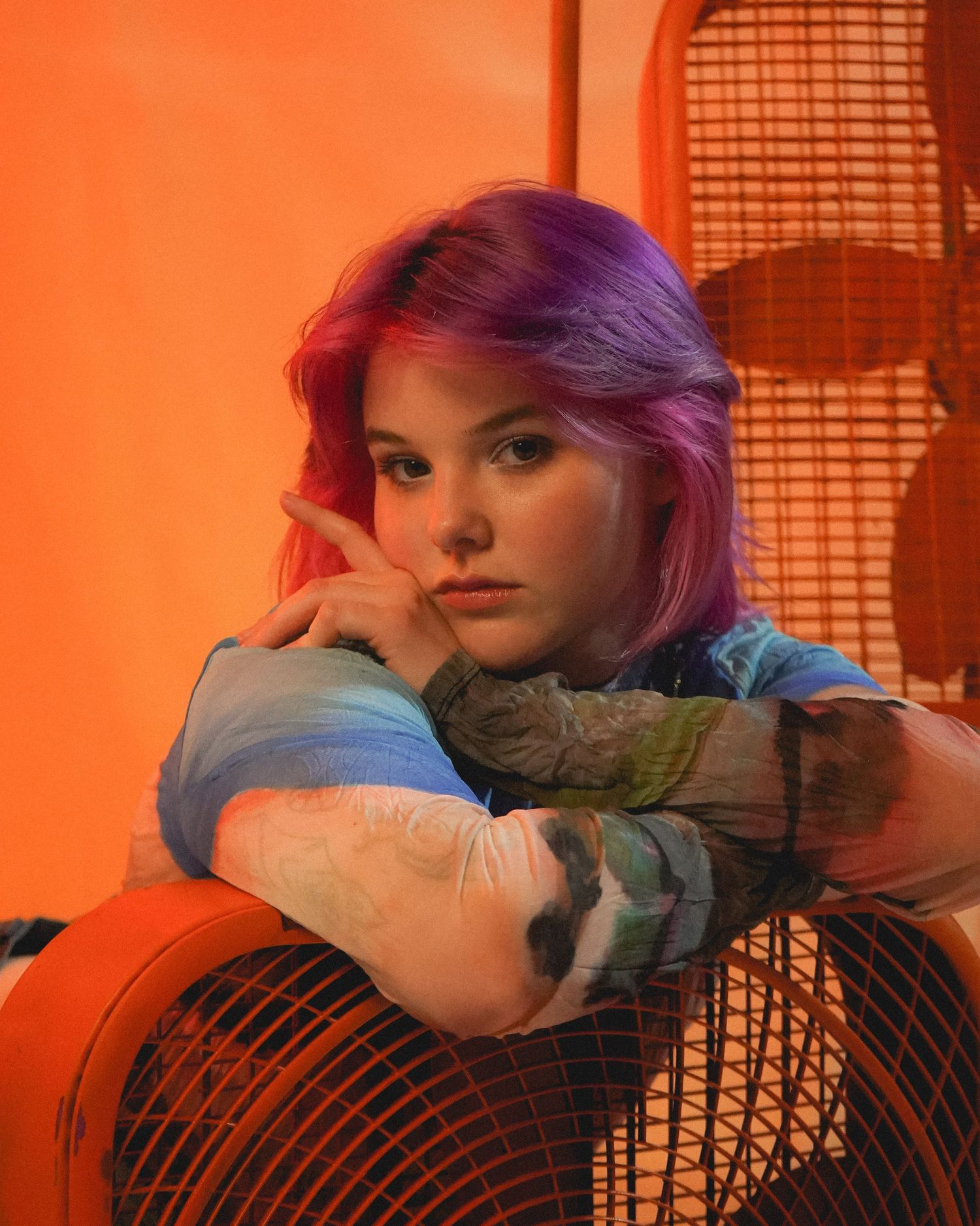 Fashion portrait with purple hair in warm orange lighting against wicker chair backdrop.