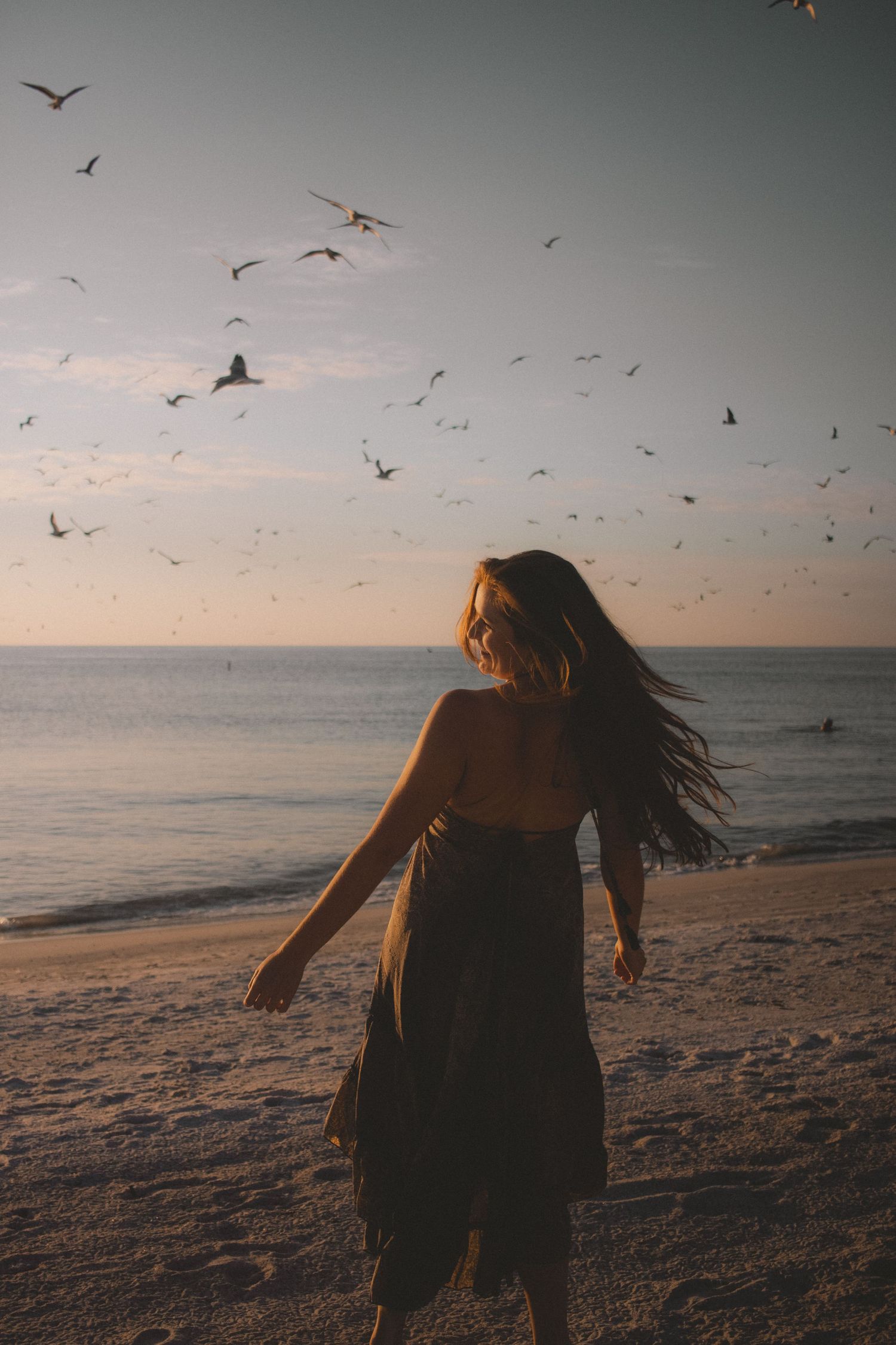 Woman in dress on beach with seagulls flying overhead at sunset.