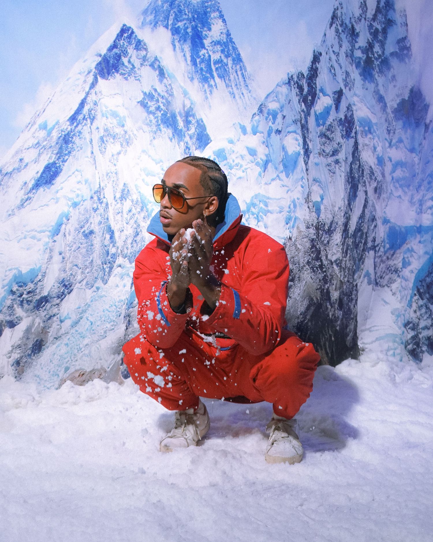 Person in red snowsuit crouches in snow with mountain peaks in background.